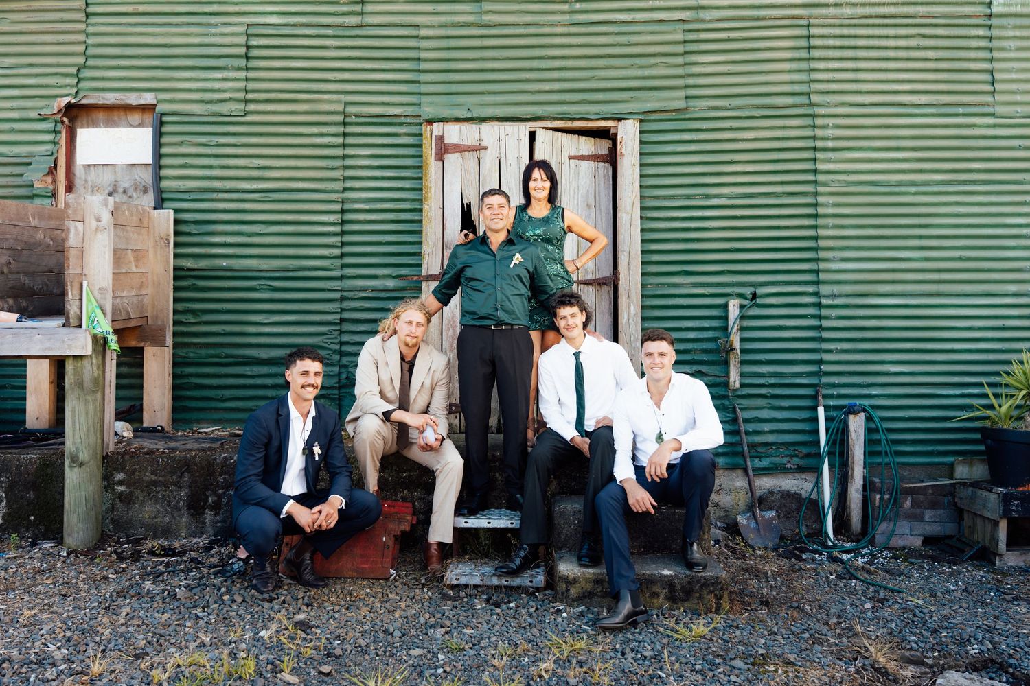 Group of six people posing in front of a rustic green corrugated iron building for a formal photo.