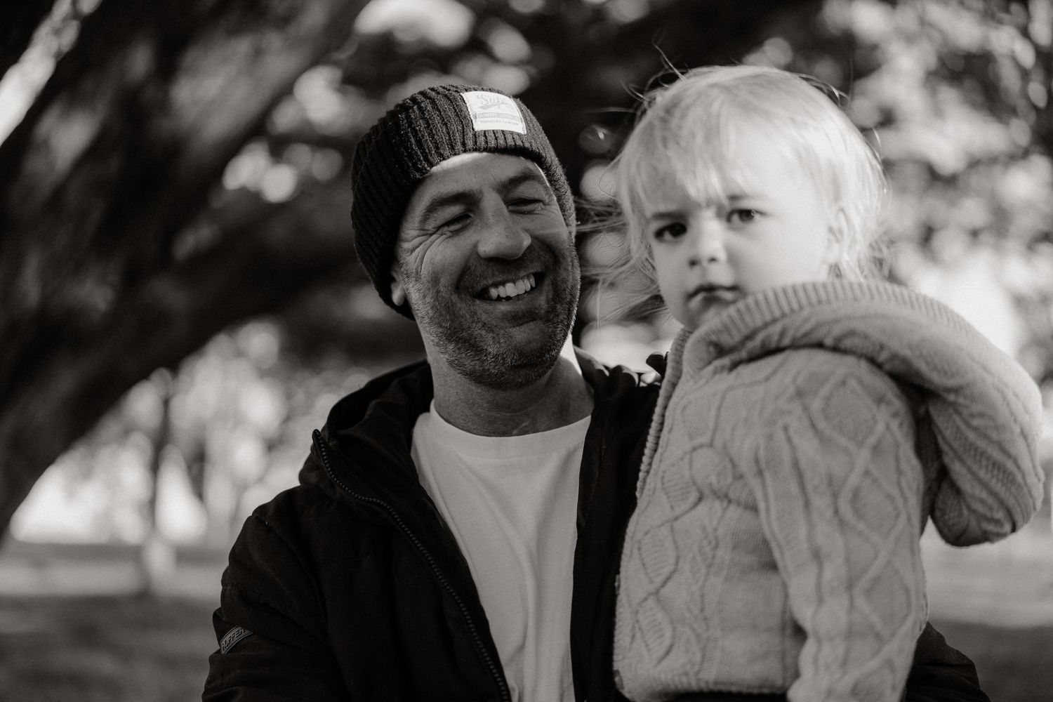 Black and white photo of a smiling man in a beanie holding a young blonde toddler outdoors.