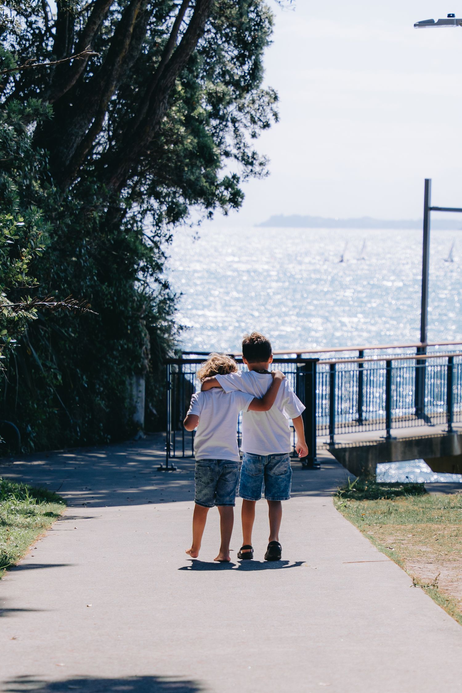 Two children walking arm in arm along a seaside path toward sparkling ocean water on a sunny day.