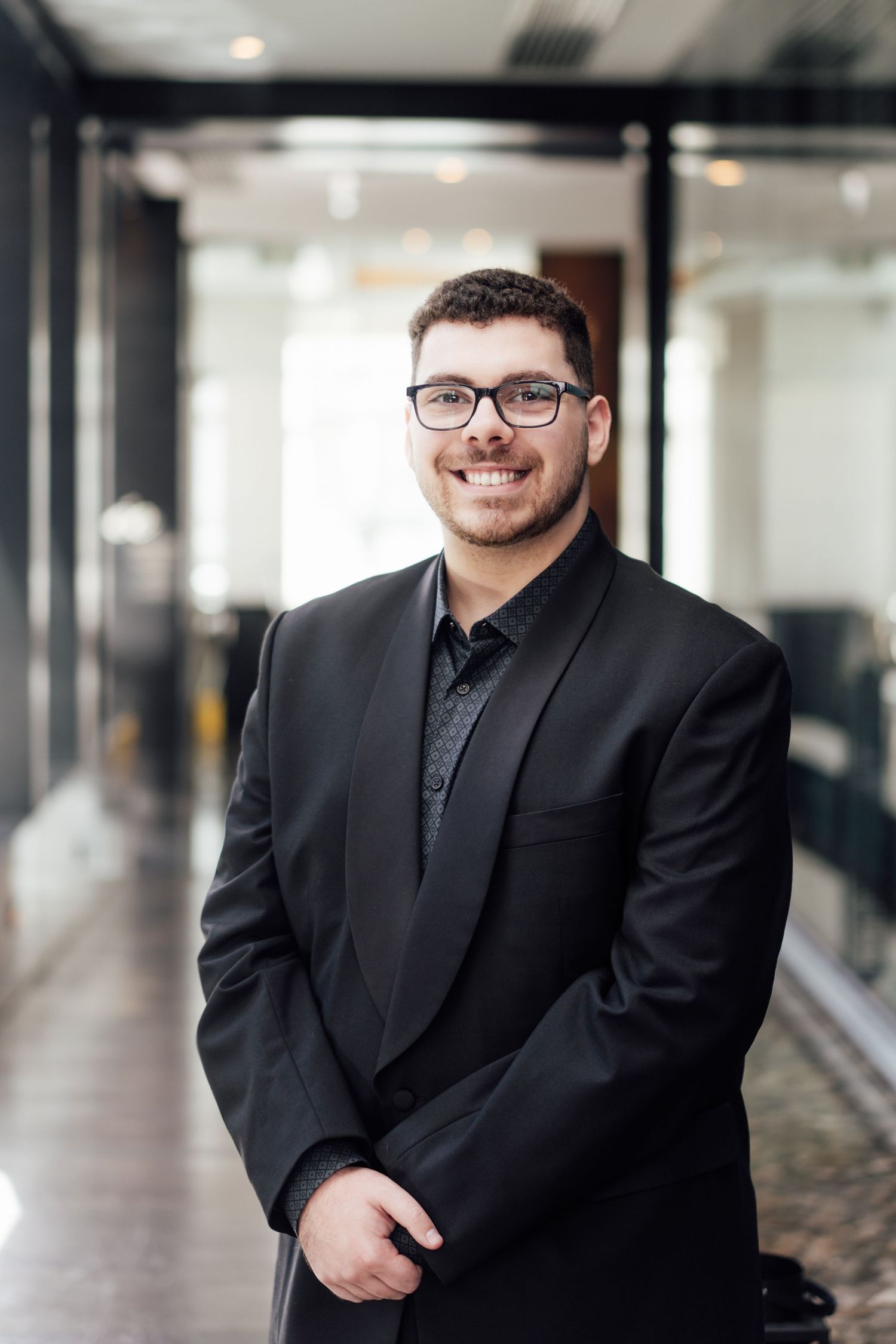 Smiling young man in black suit and glasses standing in modern office hallway for professional headshot.