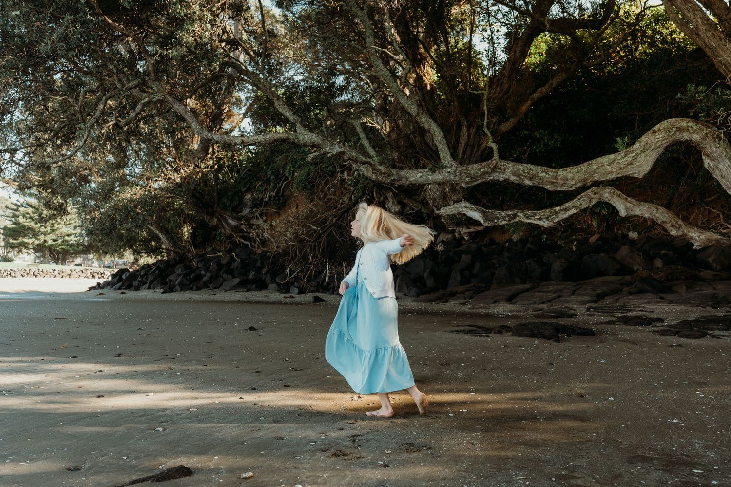 A young girl in a light blue dress twirls on a sandy beach beneath large sprawling trees.