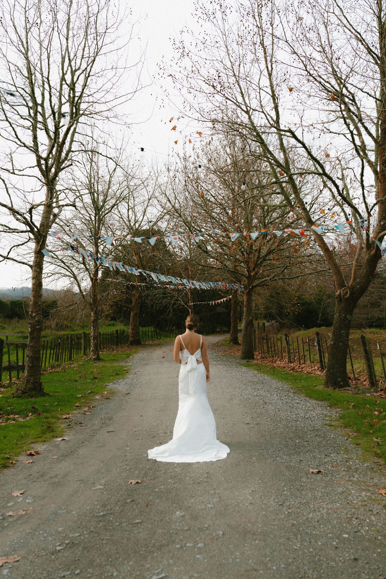 Wedding dress silhouette on tree-lined path with string lights overhead.