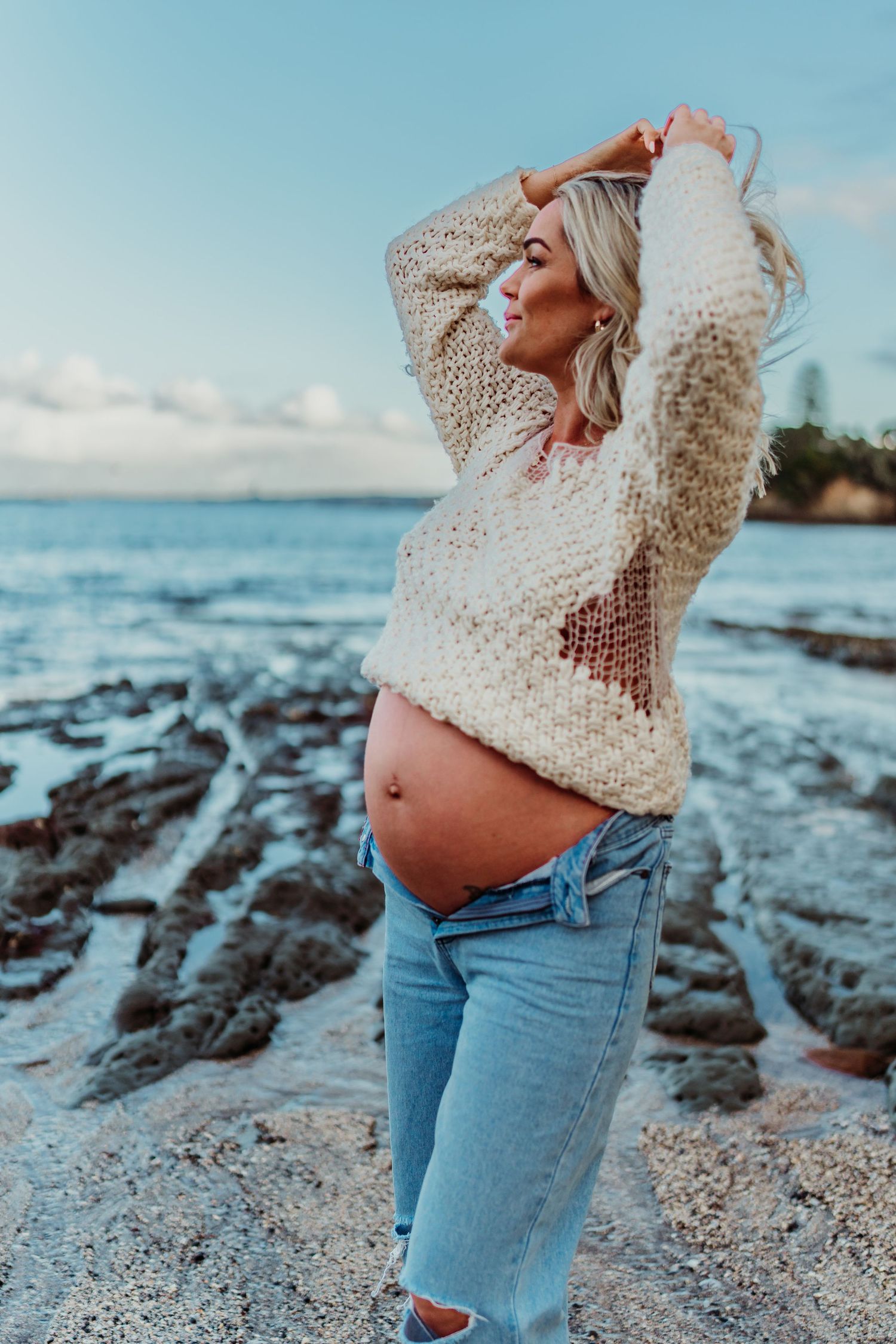 Expectant mother in cream sweater and jeans poses on rocky beach at sunset.