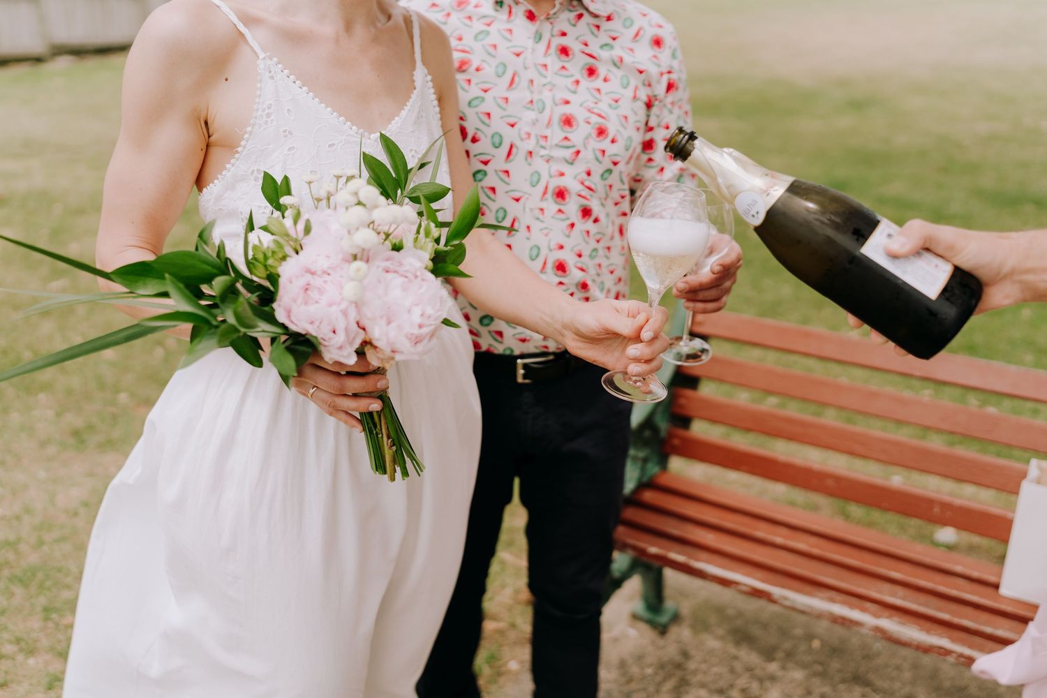 A couple holds hands while sitting on a wooden bench in a garden setting with floral decor.