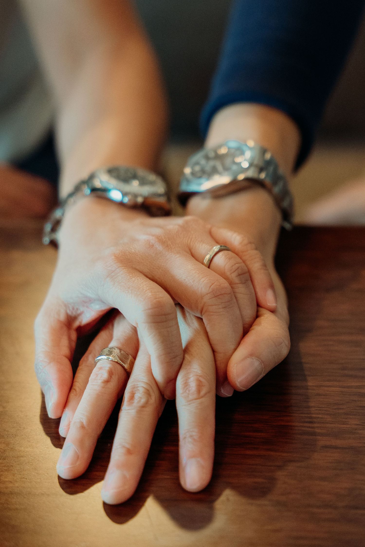 Close-up of hands wearing wedding rings resting on a wooden surface.