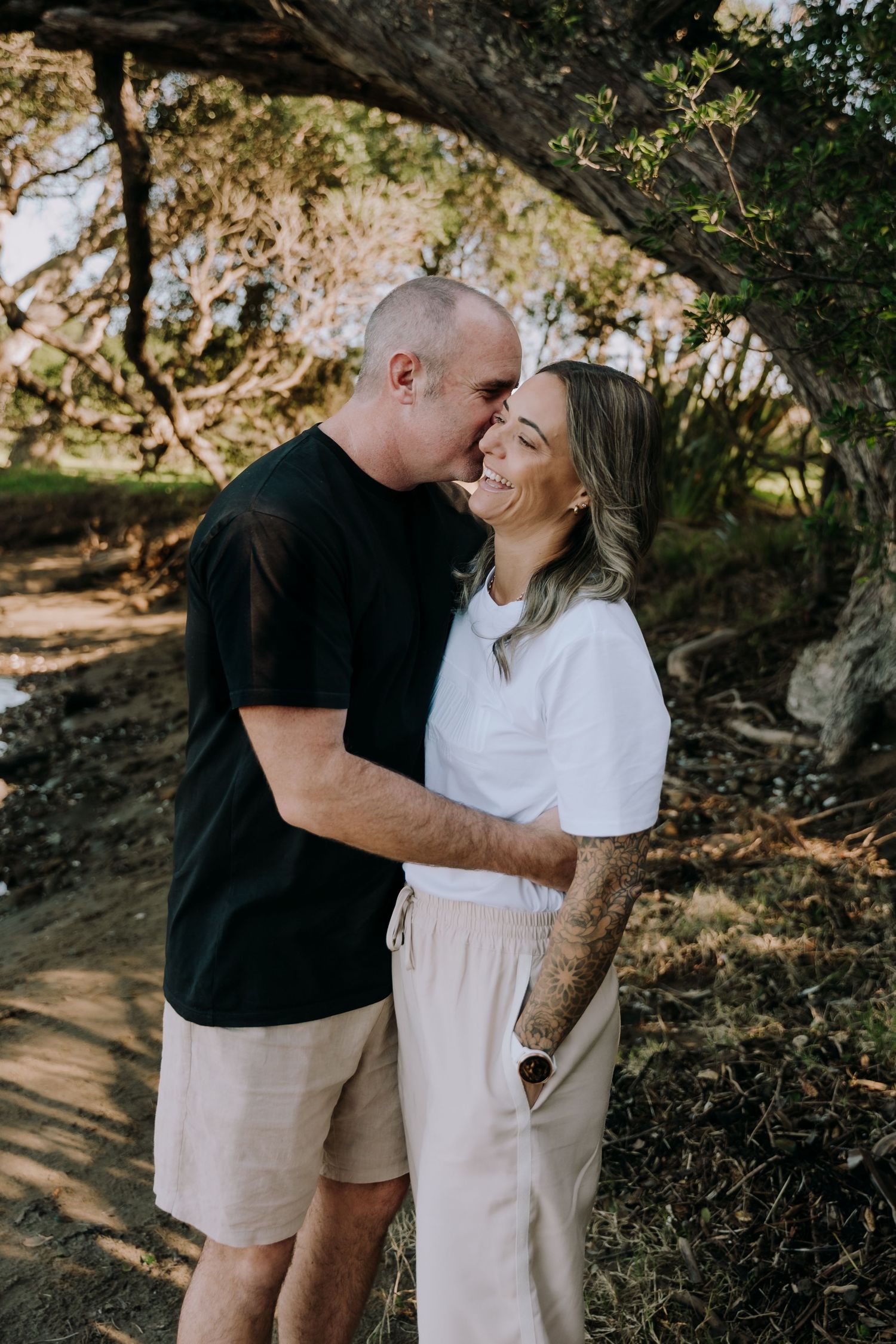 A couple shares a tender moment under a tree branch, with sunlight filtering through the leaves on a warm day.