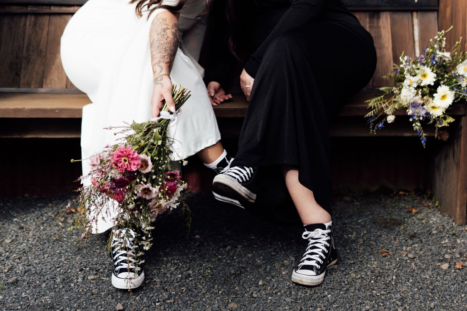 Bride and groom sitting on bench wearing black Converse sneakers, holding colorful wedding bouquets.