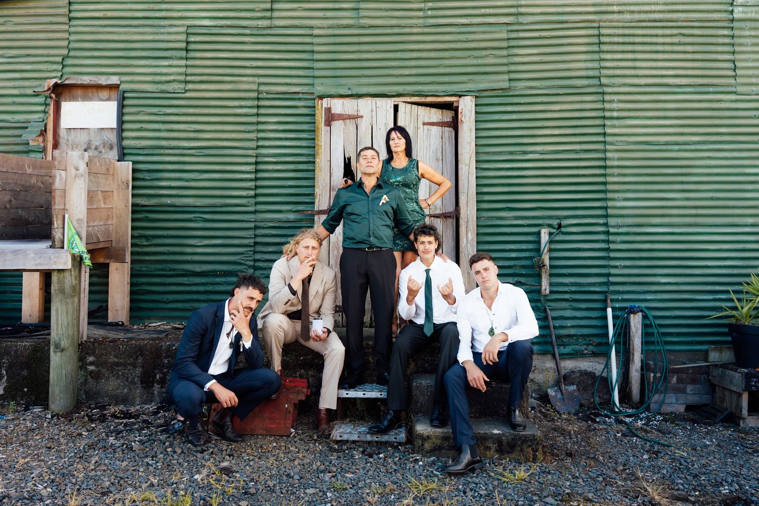 Six young adults posing casually outside a rustic green corrugated metal building with a weathered wooden door.