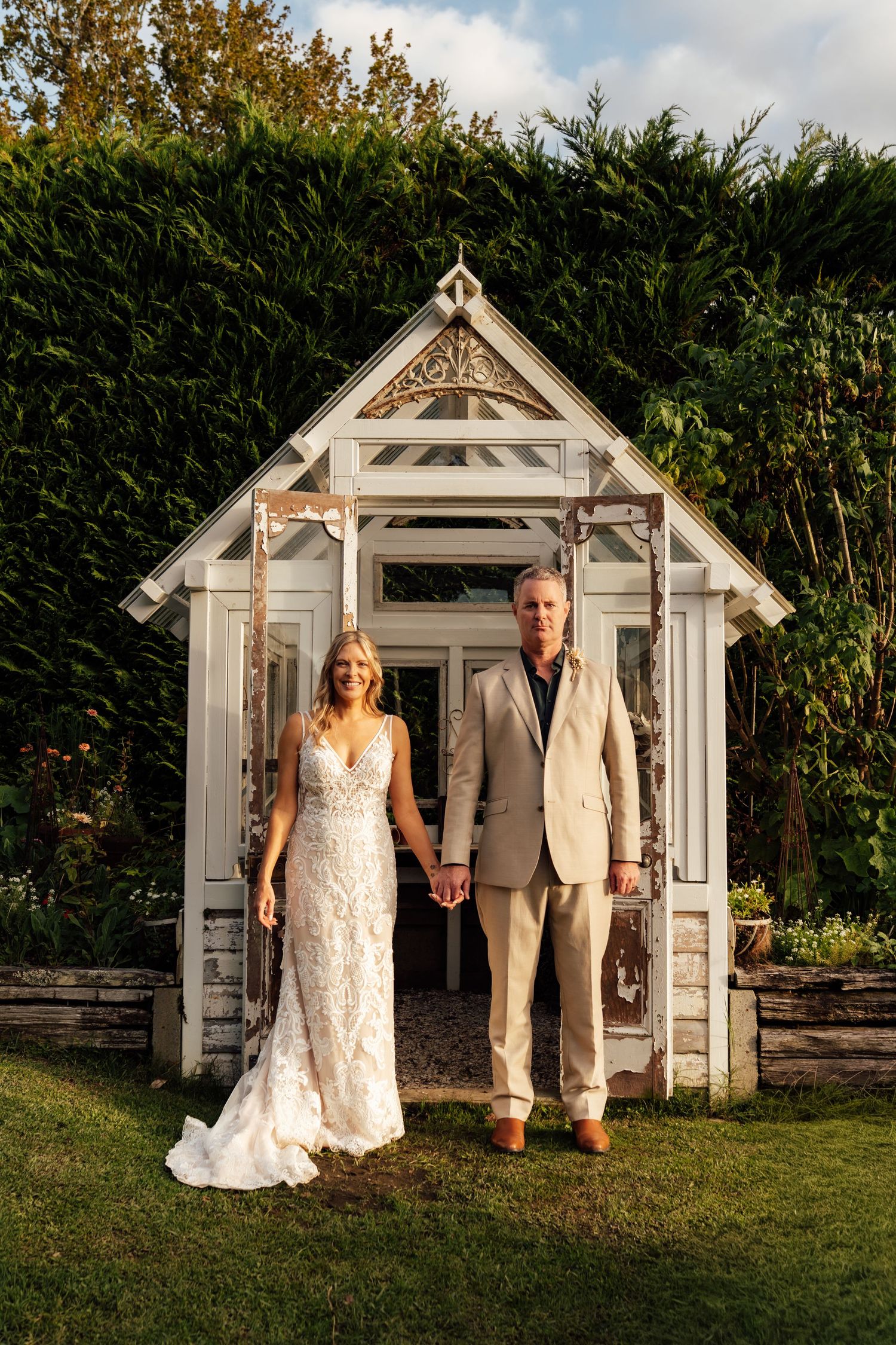 Bride in lace gown and groom in tan suit hold hands before a white garden gazebo surrounded by lush greenery.
