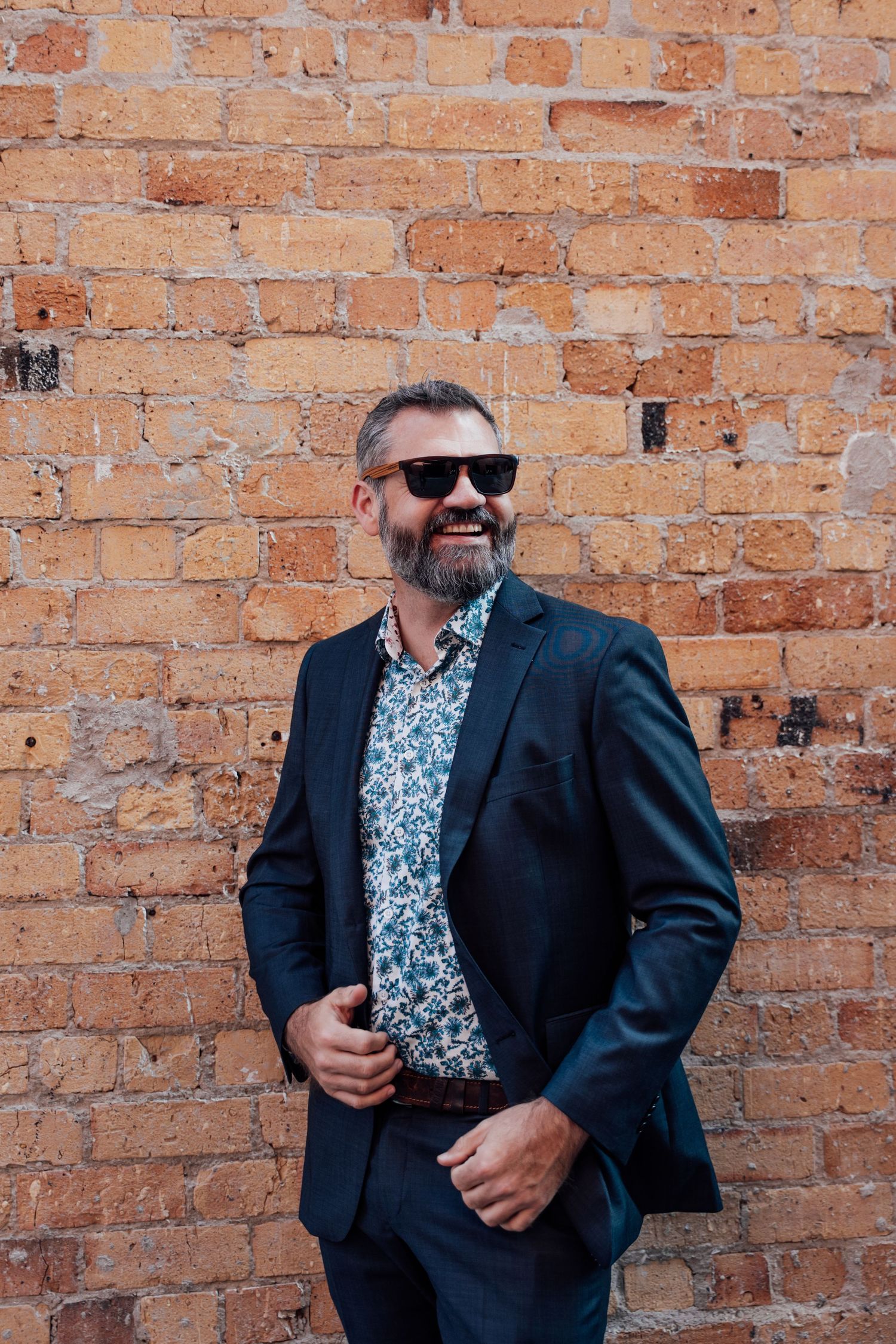 Smiling bearded man in sunglasses, floral shirt, and dark blazer posing against a rustic brick wall.