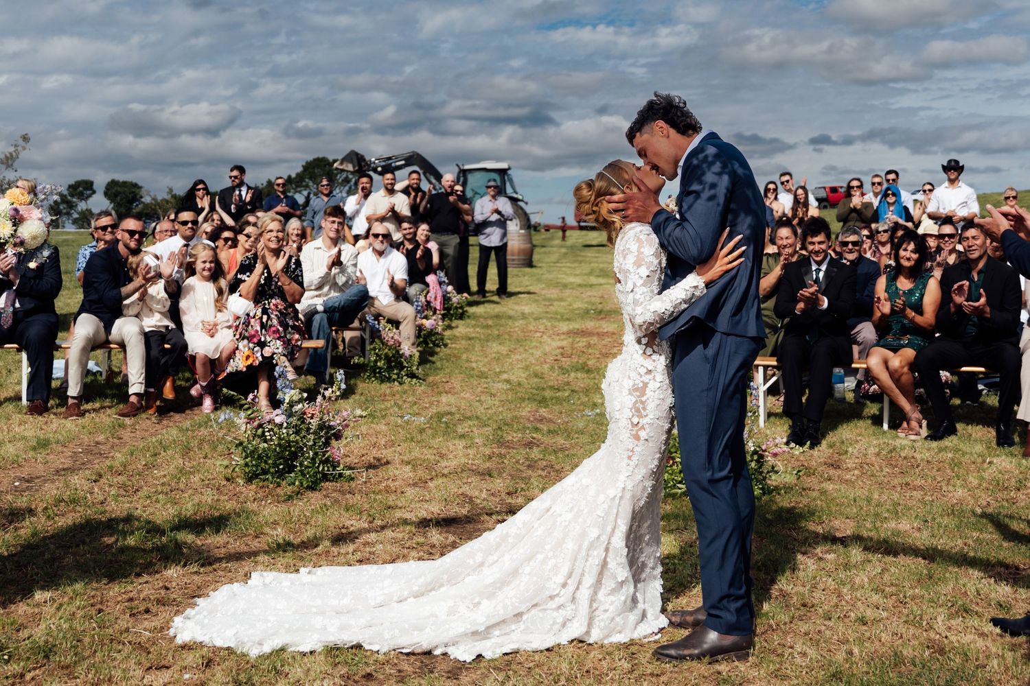 Couple sharing first kiss in white lace gown and blue suit at outdoor wedding ceremony with guests seated on grass.