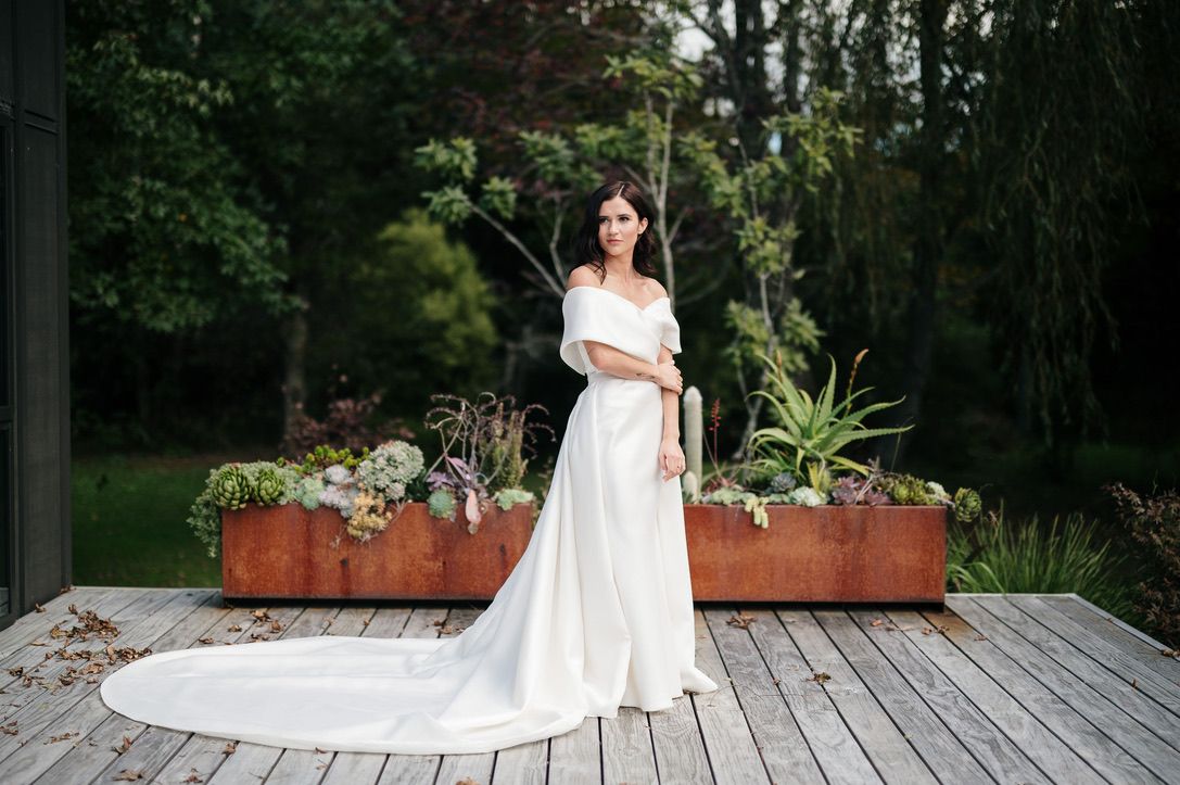 A bride in a white wedding dress poses elegantly on an outdoor wooden deck surrounded by greenery.