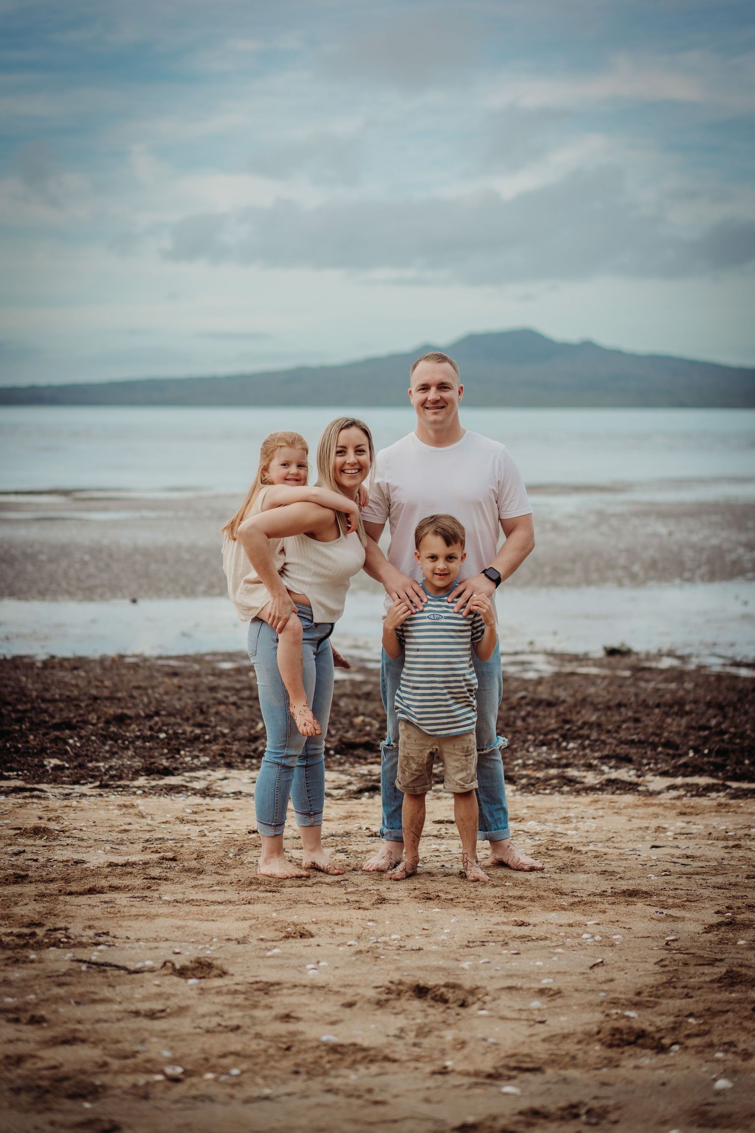 A family enjoys a casual moment together on a rocky beach with mountains visible in the distance.