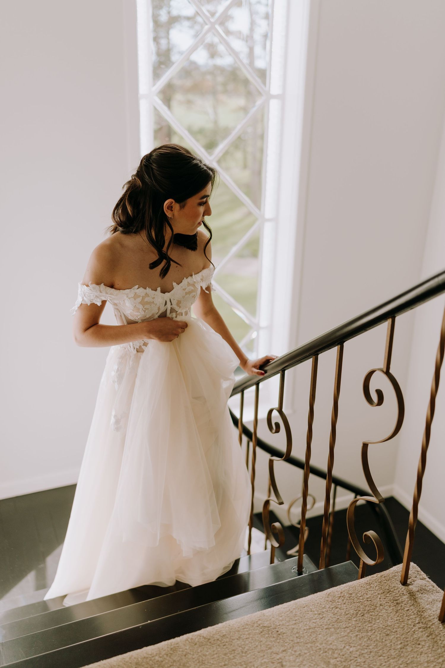 A bride in a white wedding gown gracefully ascends a curved staircase with an ornate iron railing.