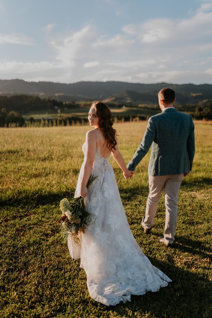 A couple walks through a golden field at sunset with mountains in the background during their wedding photoshoot.