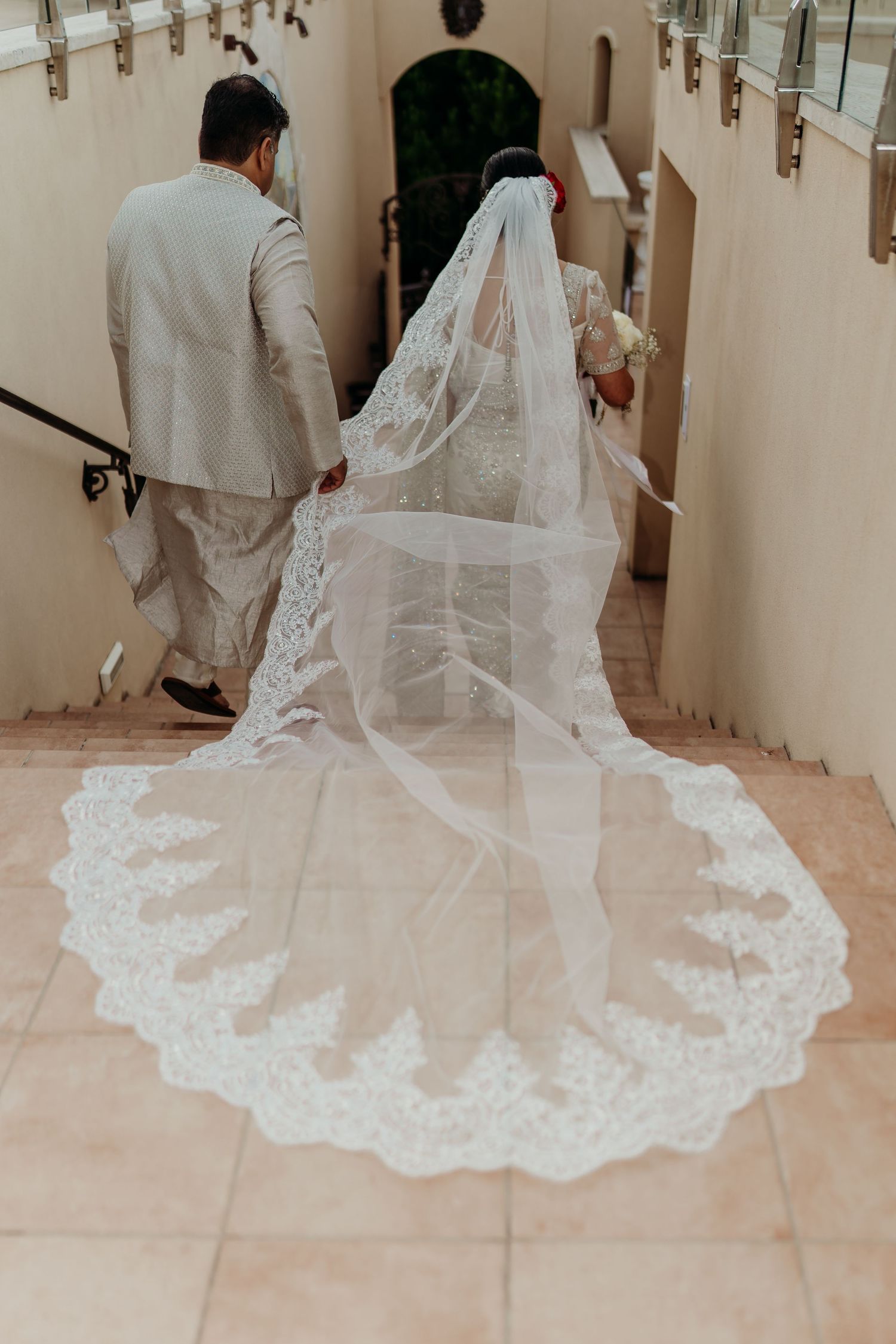 Bride in lace wedding gown with long cathedral veil walks up stairs with groom in beige suit.