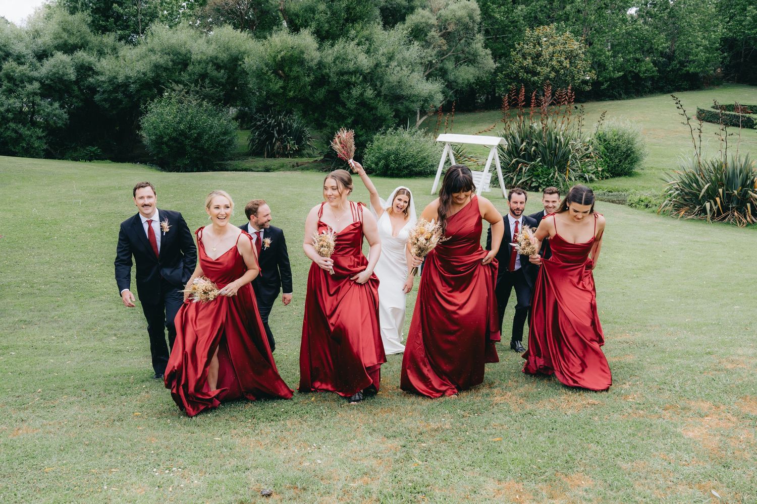 Bridal party in red satin dresses celebrating on green lawn with bride in white gown and groomsmen in dark suits.