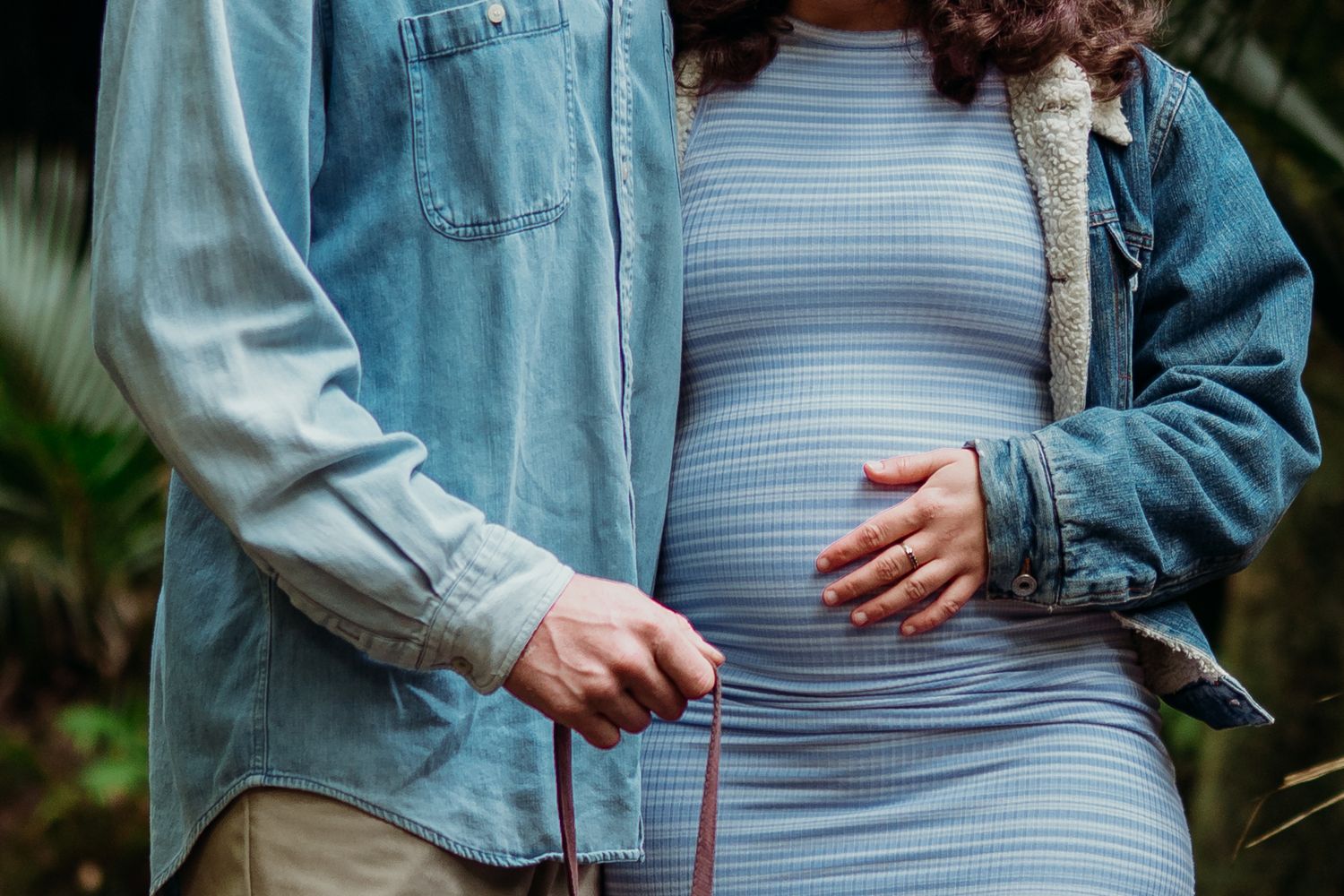 Pregnant woman in blue dress with partner outdoors, both touching baby bump, maternity couple photo.
