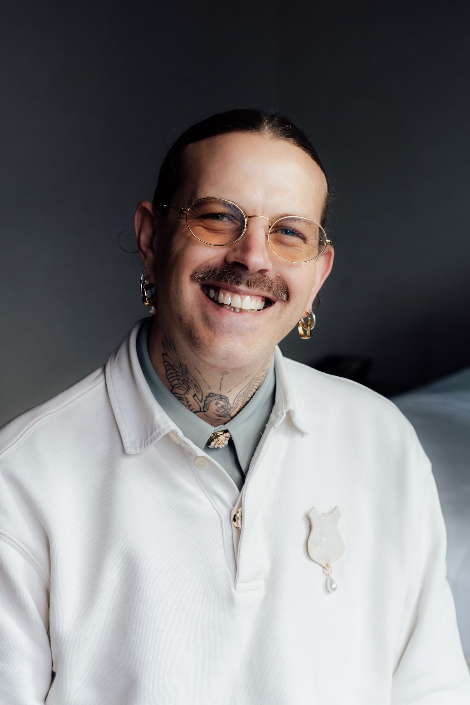 Smiling person with mustache, tattoos, gold hoop earrings, tinted glasses, and white polo shirt with pin badge.