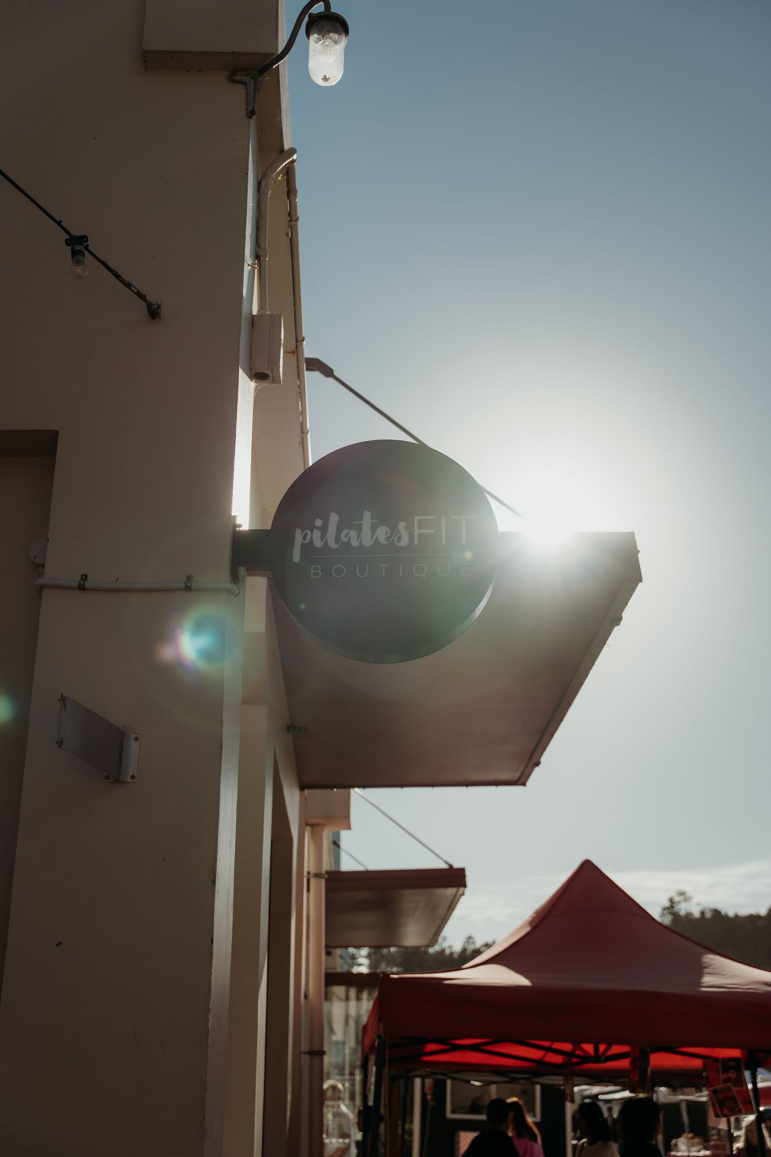 A circular Pilates studio sign mounted on a building exterior with sun flare and red market tents visible below.