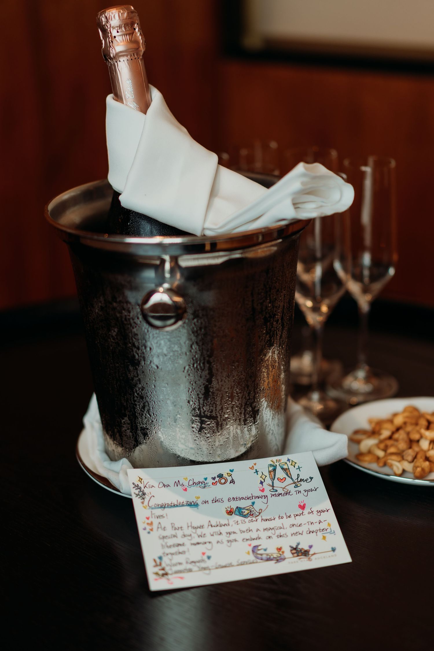 Champagne bottle chilling in an ice bucket with a handwritten note and wine glasses on a dark wooden table.