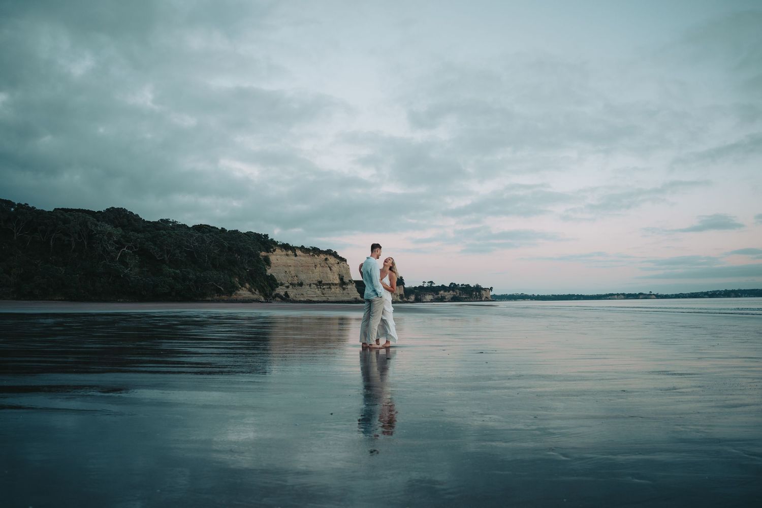 A couple walks along a misty beach shoreline at dusk with mountains and cloudy skies in the background.
