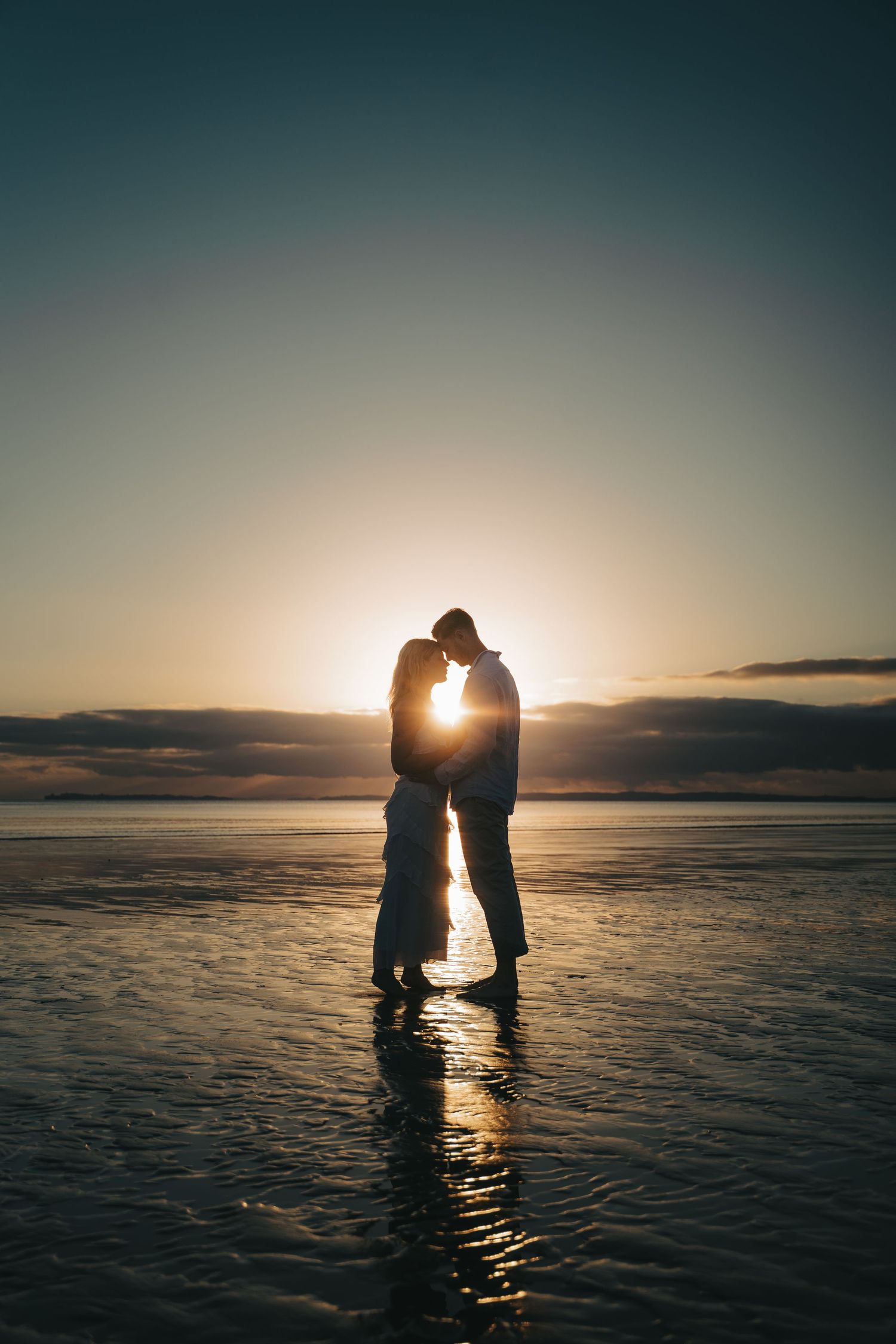 Romantic couple silhouetted against golden sunset on a reflective beach, sharing an intimate moment by the ocean.