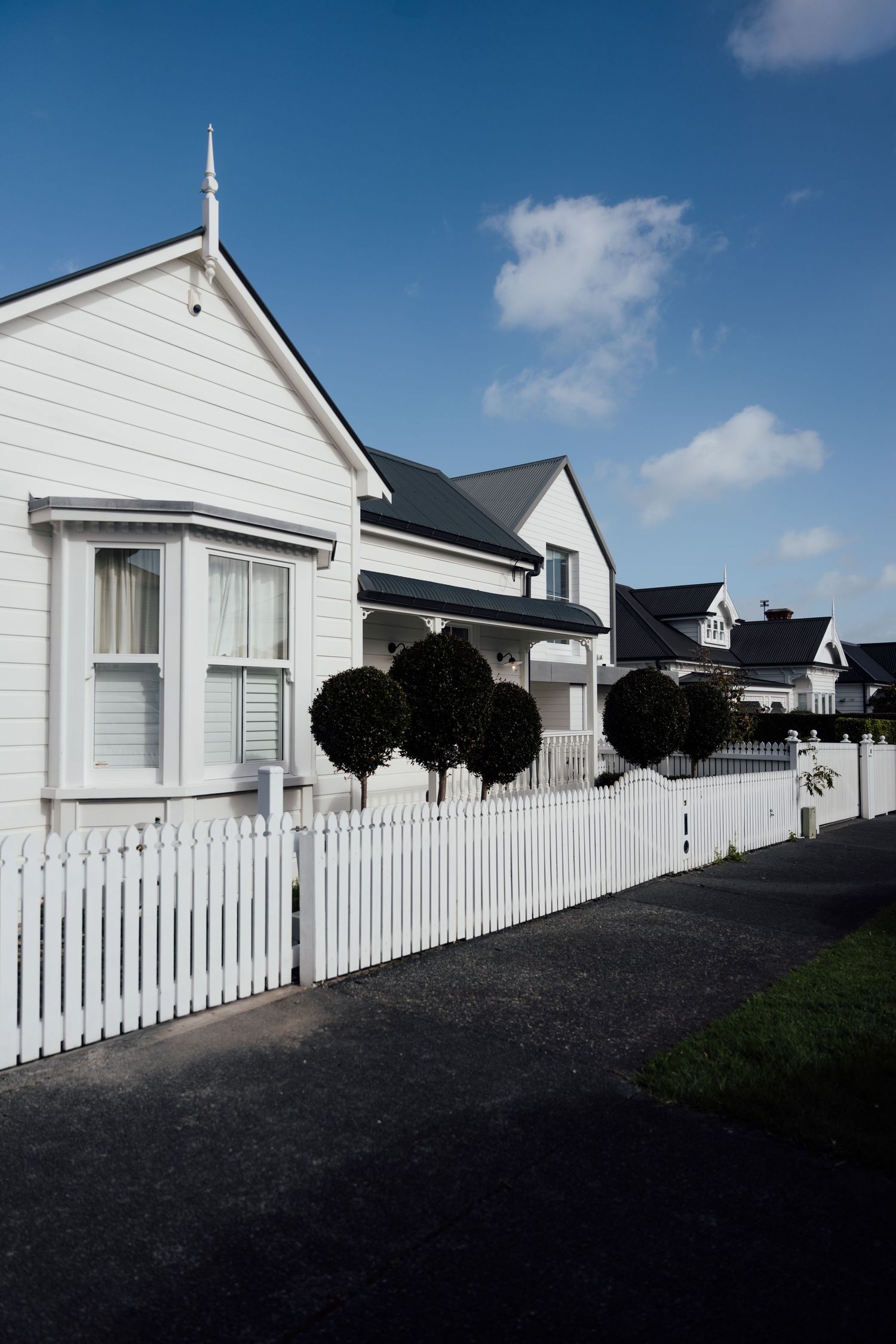 White weatherboard houses with picket fence and topiary trees under blue sky in a classic New Zealand suburban street.