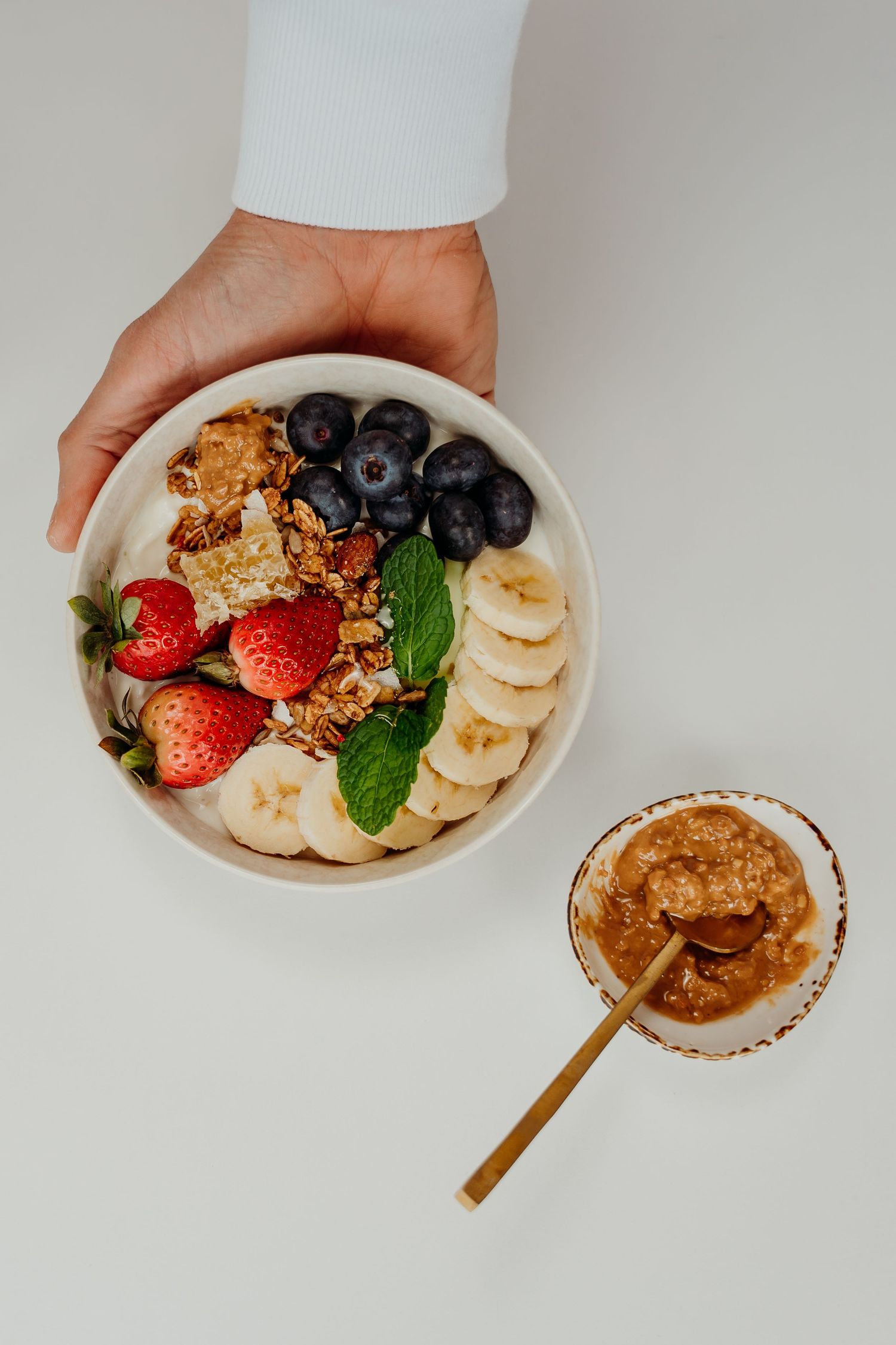 Overhead view of a hand holding a bowl of granola topped with strawberries, blueberries, and banana slices.