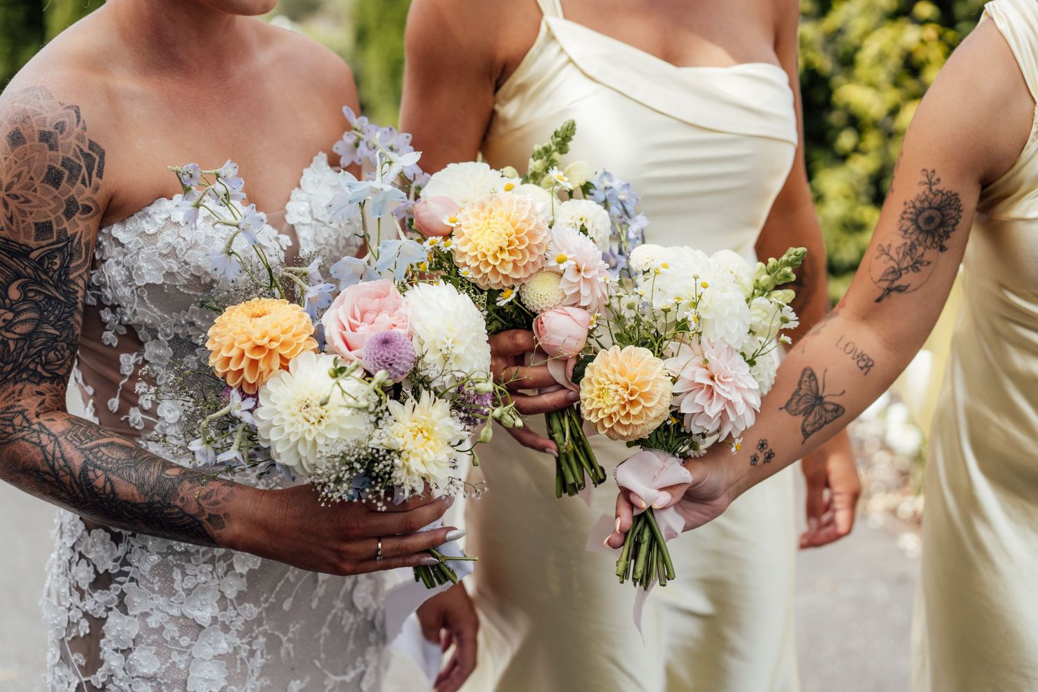 Bride and bridesmaids holding pastel bouquets of dahlias and roses, wearing white gowns with floral tattoos visible.