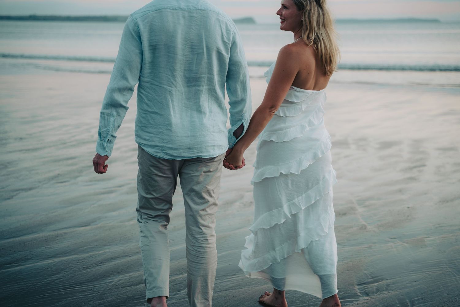 Couple holding hands walking on a beach at sunset, wearing white outfits, with ocean waves in the background.