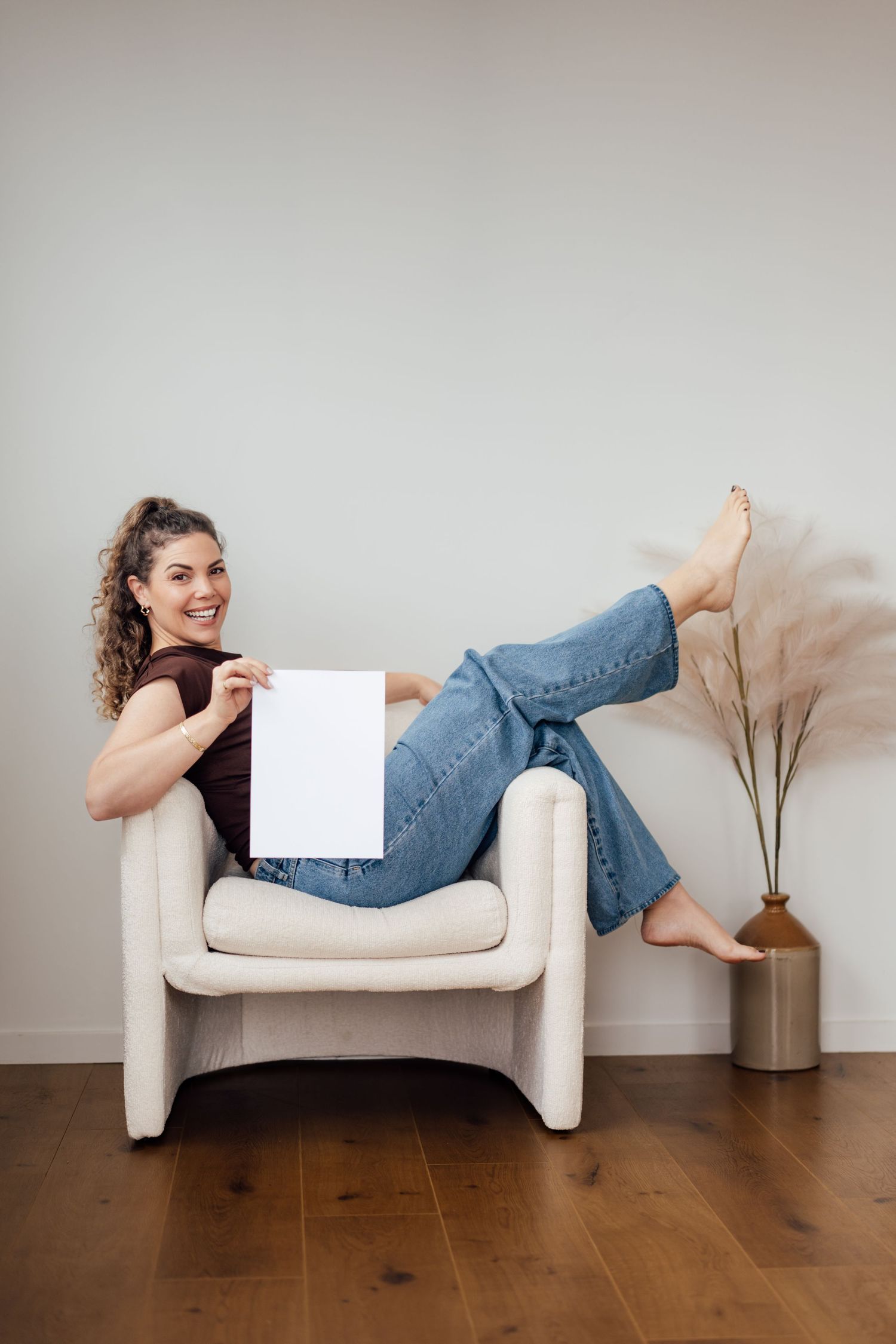 Smiling woman in jeans holding blank white paper while relaxing in a modern armchair on hardwood floor.