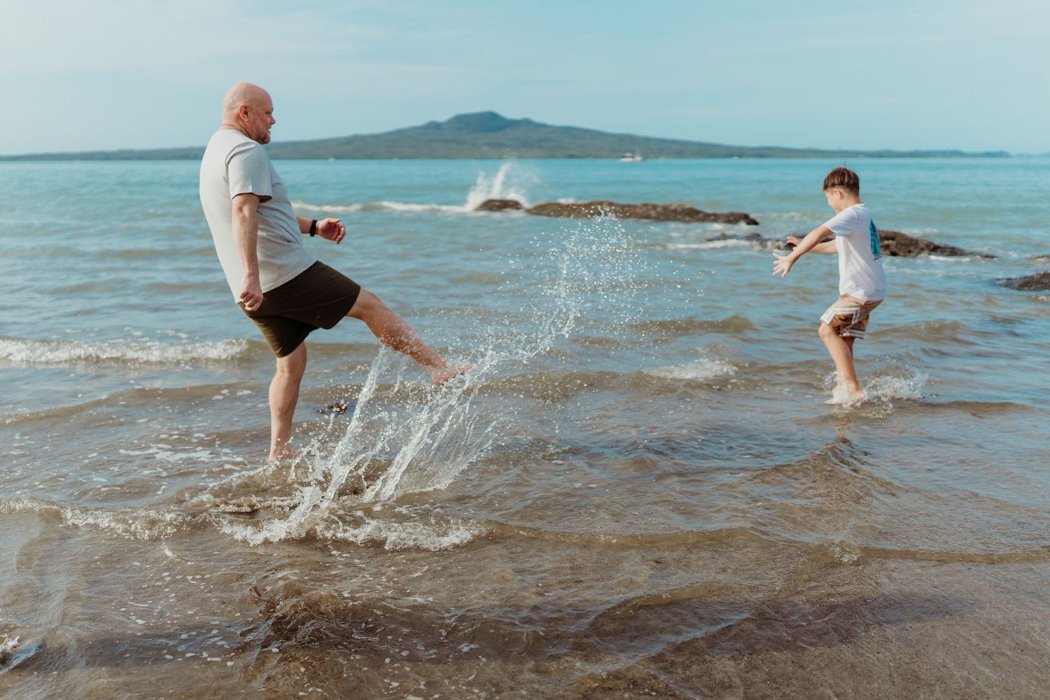 People playing and splashing in shallow ocean water at the beach with a distant island visible in the background.
