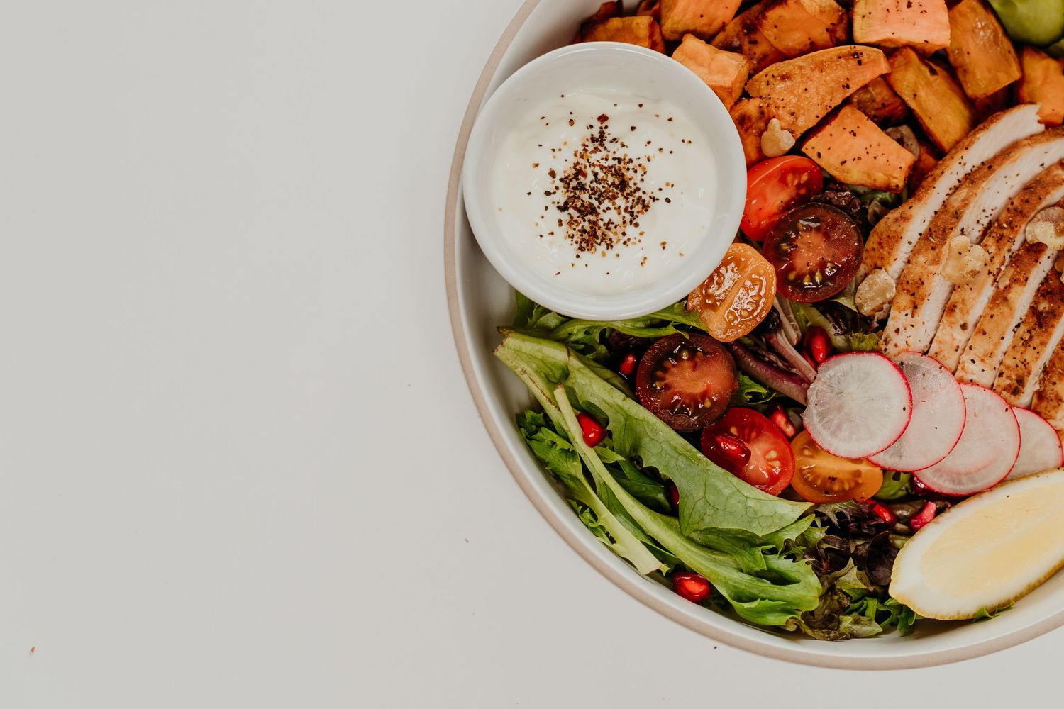 Colorful grain bowl with grilled chicken, roasted sweet potatoes, fresh salad greens, tomatoes, radishes, avocado, and creamy dressing.
