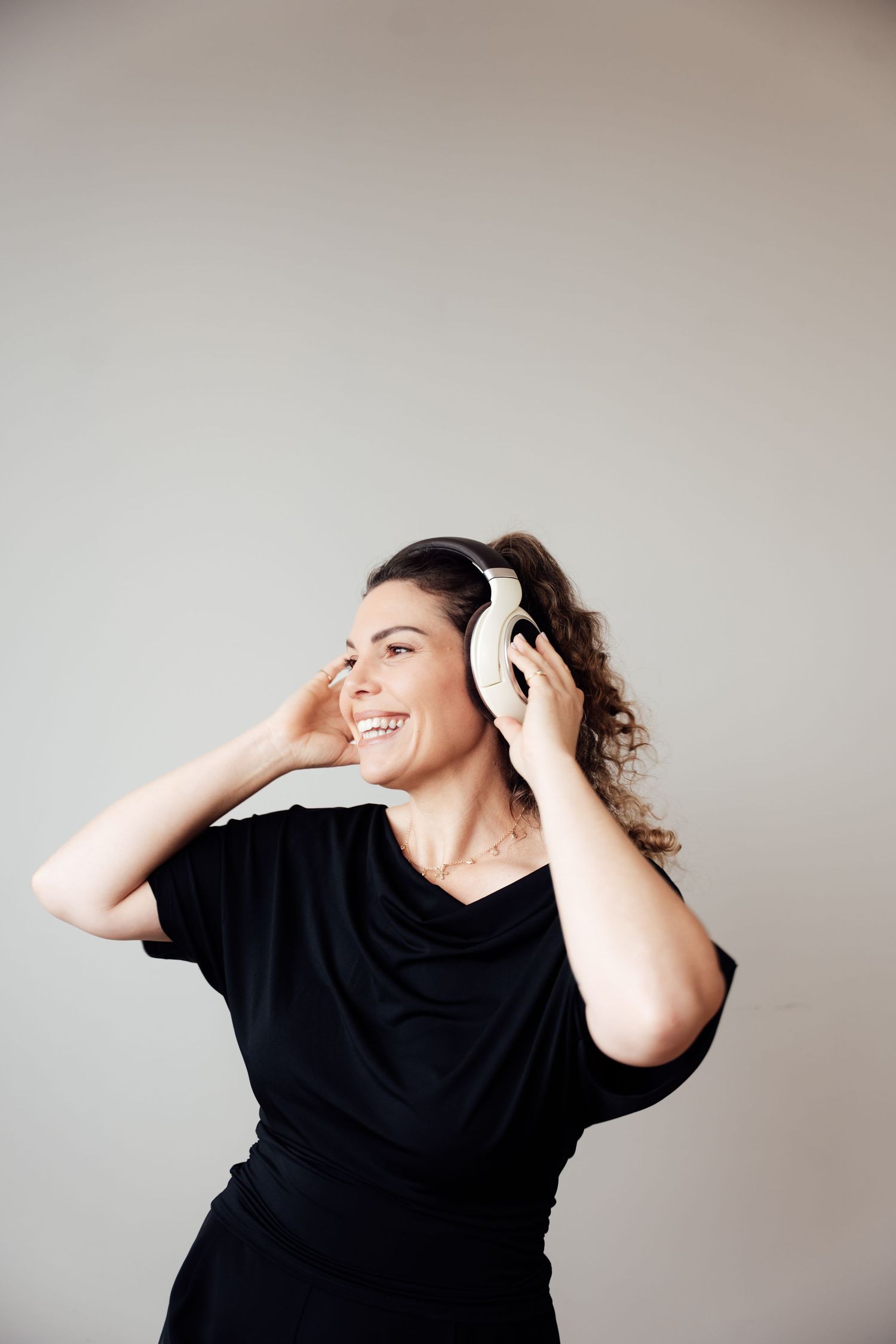 Smiling woman in black dress wearing white headphones, looking sideways, listening to music against gray background.