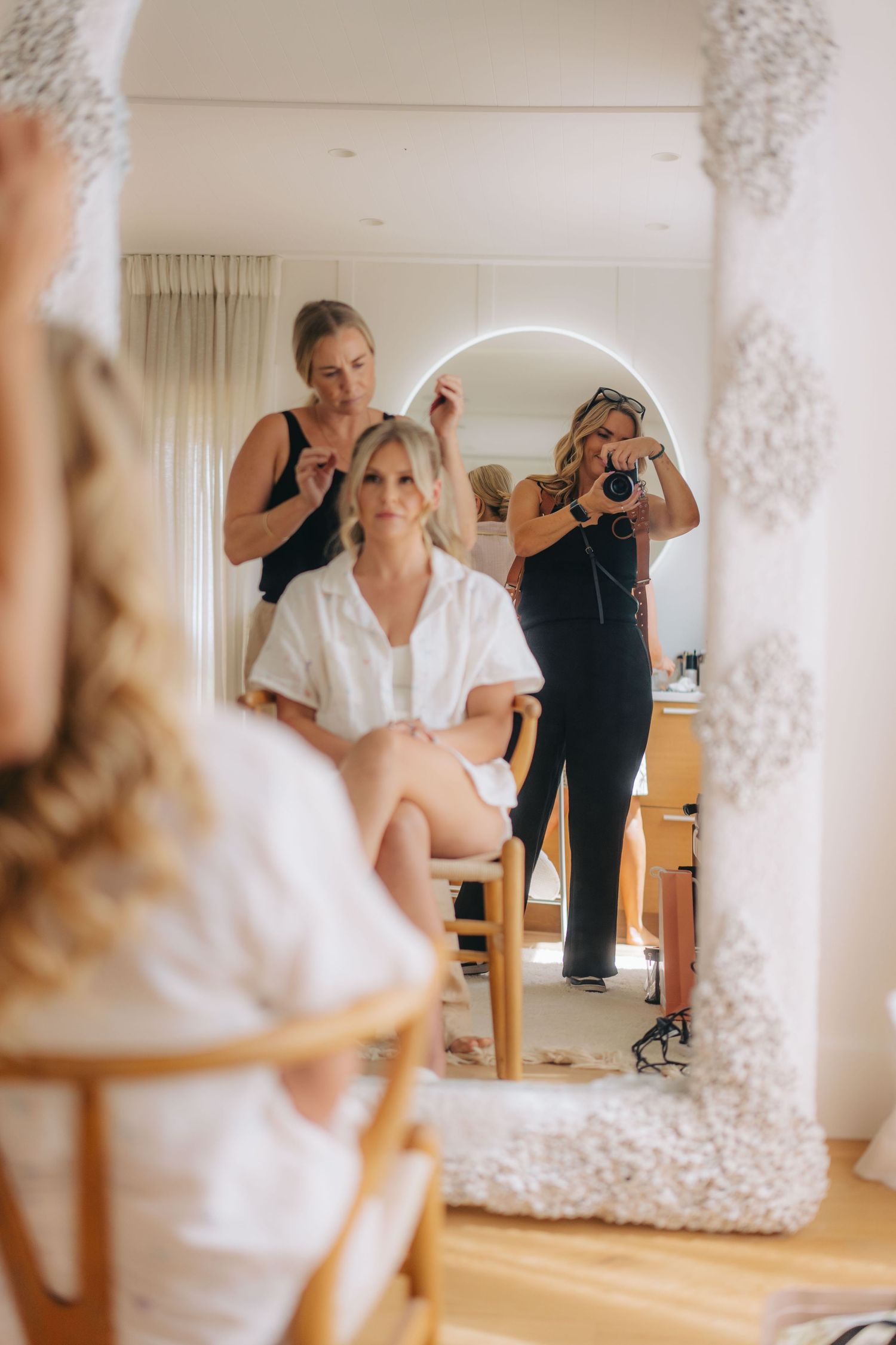 A stylist works on a bride's hair in a bright white room during wedding preparations.