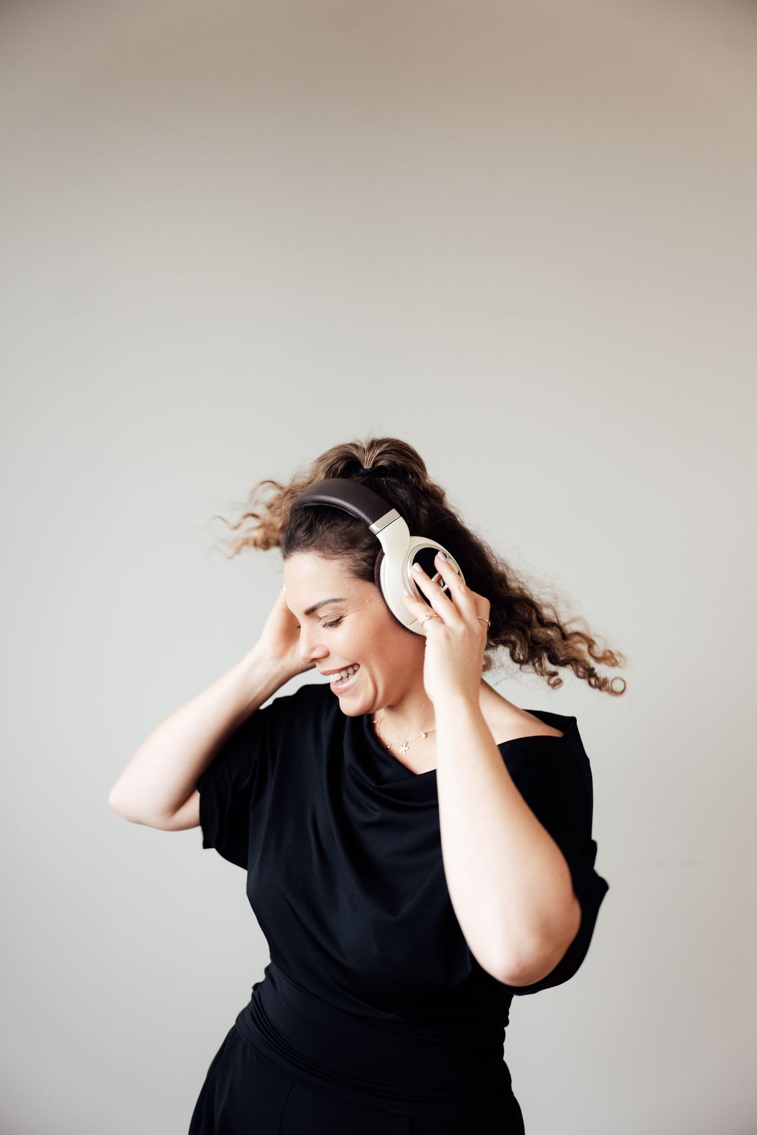 Happy woman in black dress dancing with white headphones, curly hair flowing, eyes closed, enjoying music.