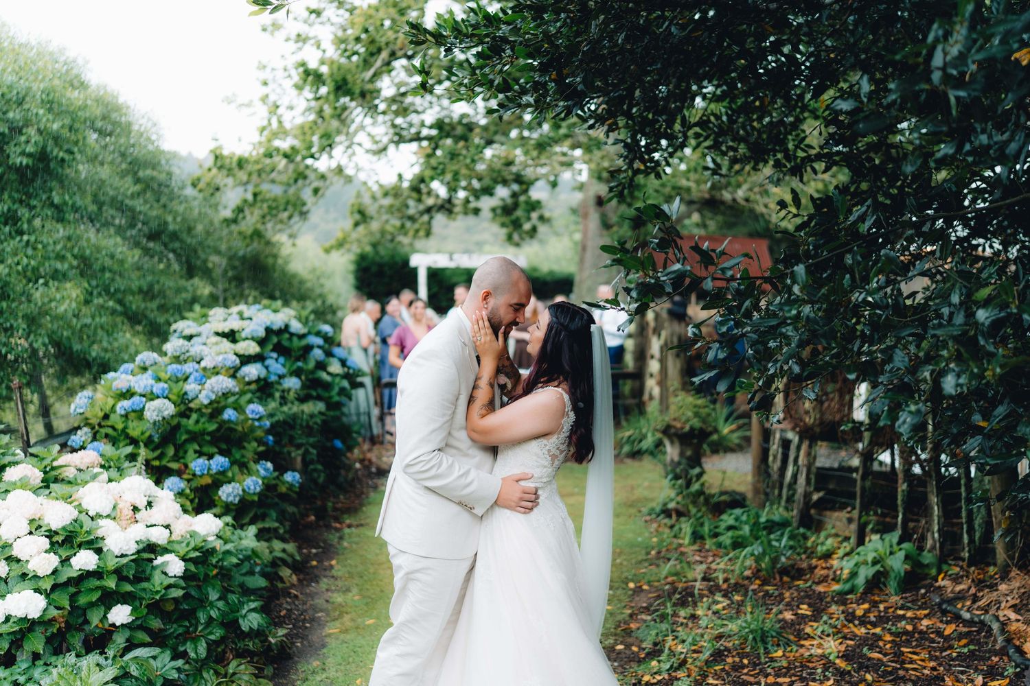A romantic wedding couple embraces in a lush garden setting surrounded by vibrant summer flowers and greenery.