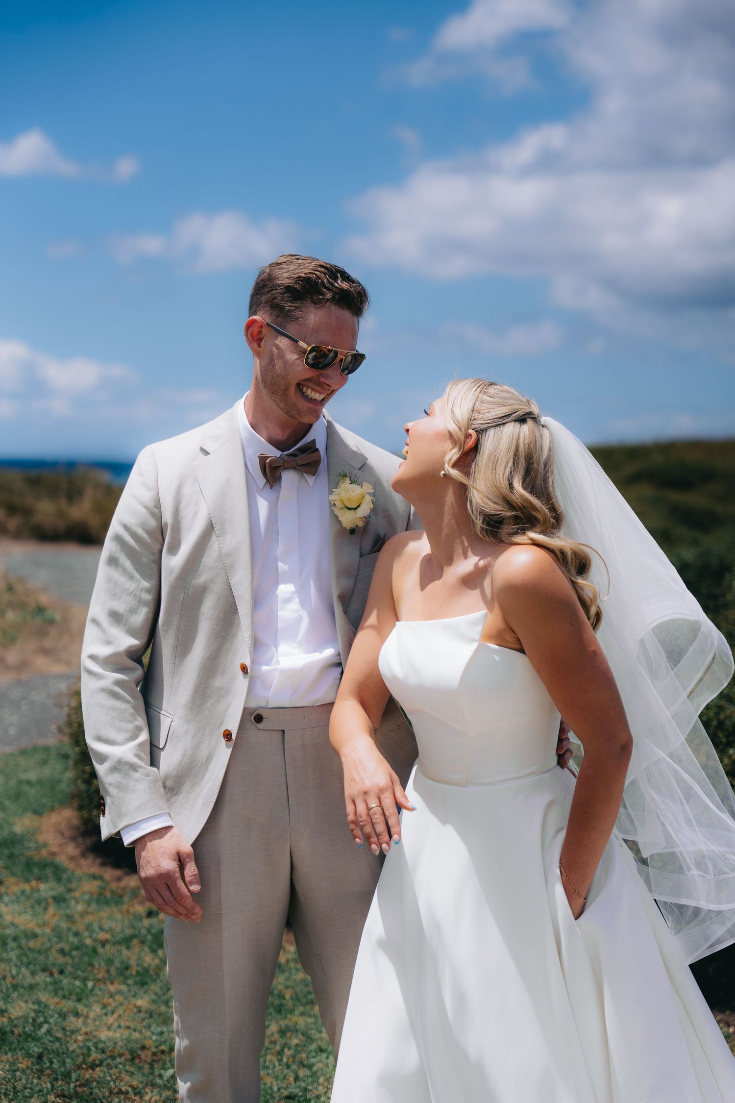 A smiling bride and groom in wedding attire share a joyful moment outdoors under a bright blue sky.
