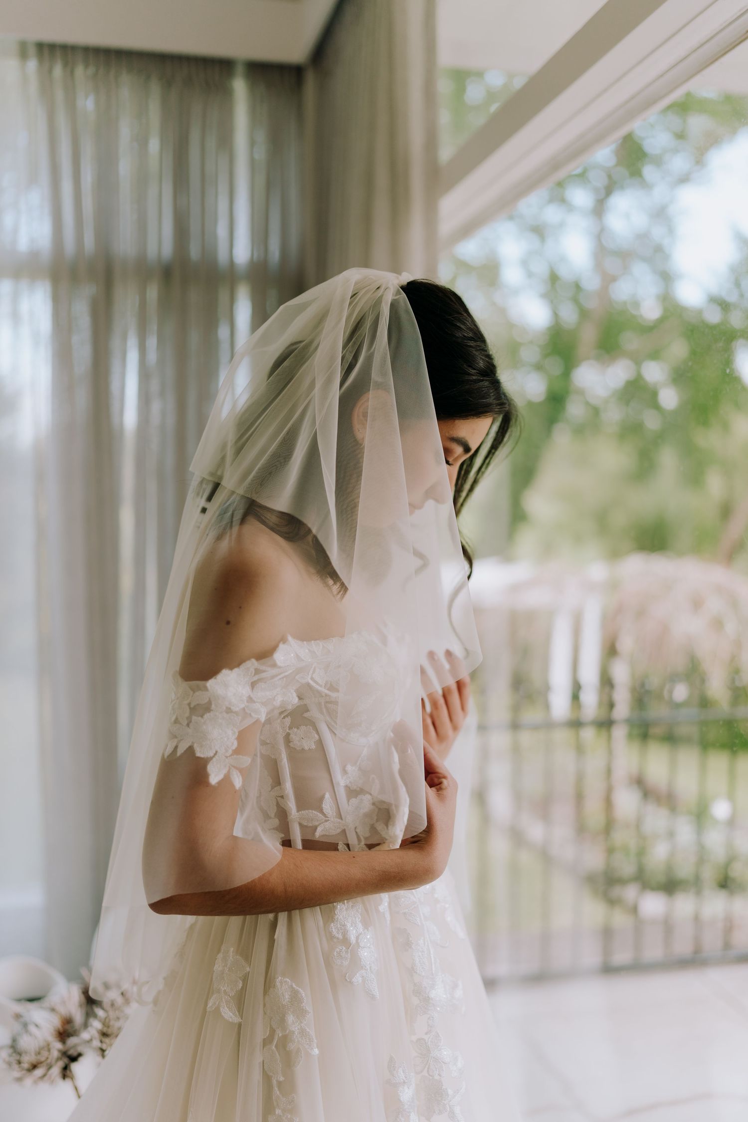 A bride in a delicate white lace wedding dress and veil stands by a window in soft natural light.
