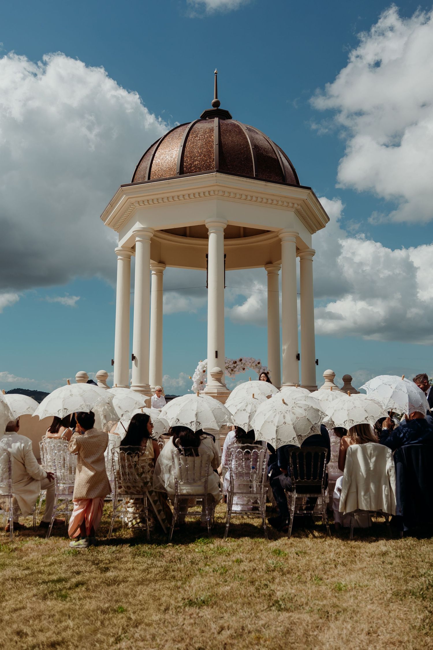 Outdoor wedding ceremony under a white columned gazebo with copper dome, guests seated with white parasols under blue sky.