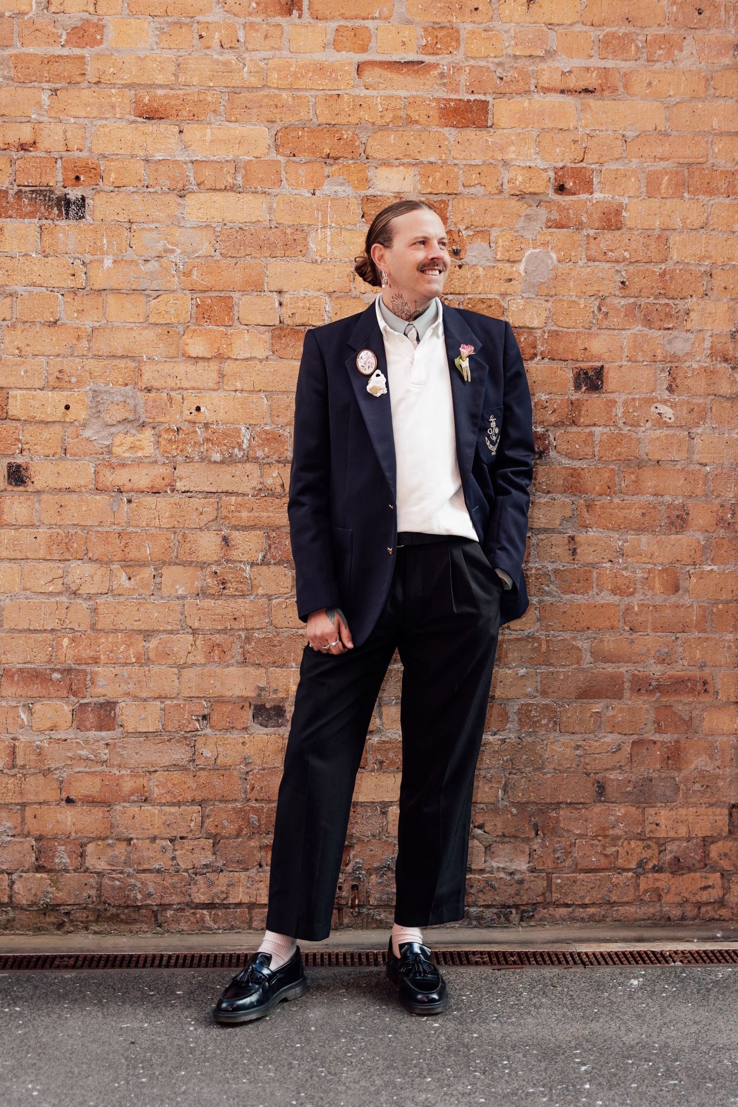 Man in black suit with pins smiling against brick wall.