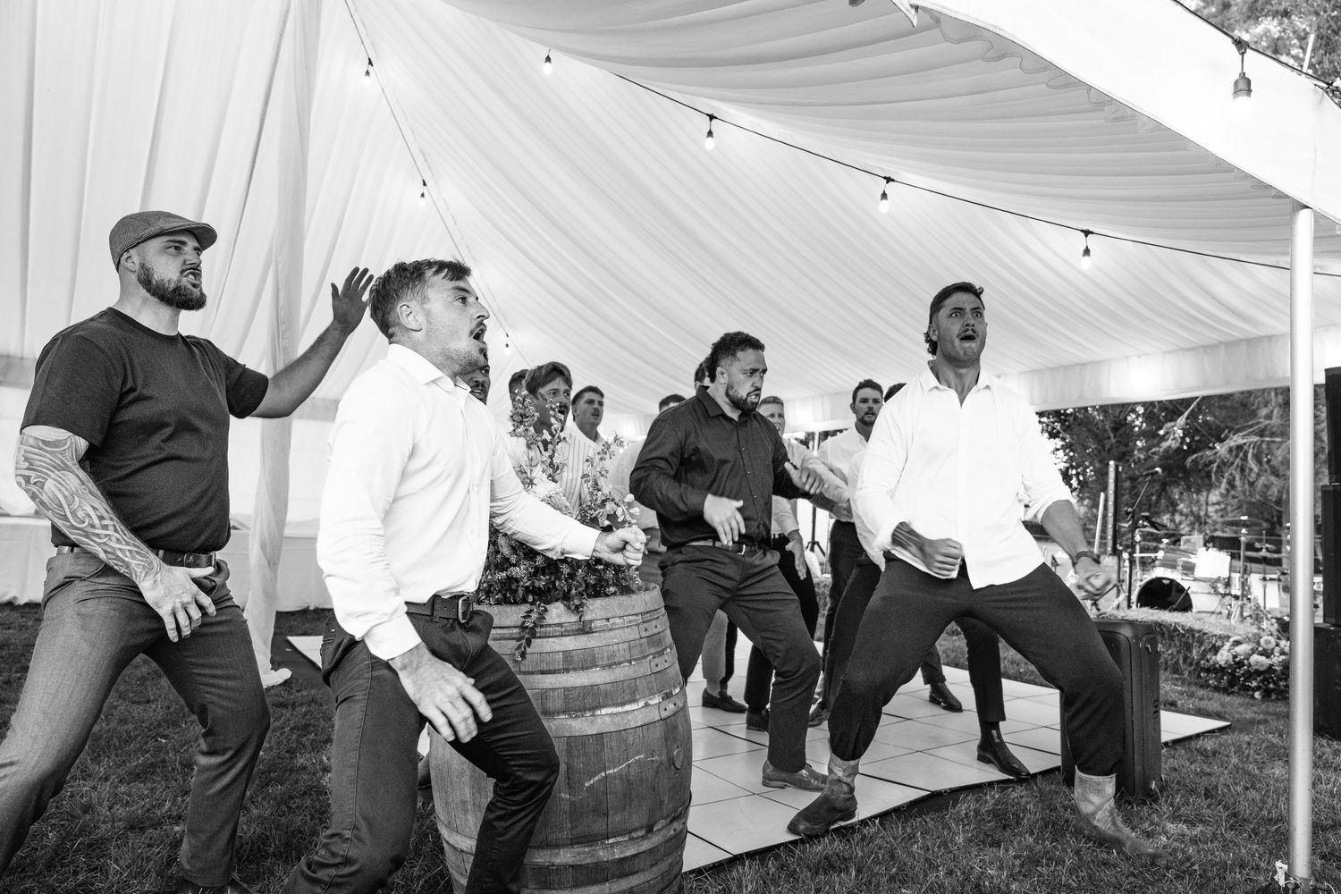 Black and white photo of groomsmen performing a lively haka dance under a marquee at a wedding reception.