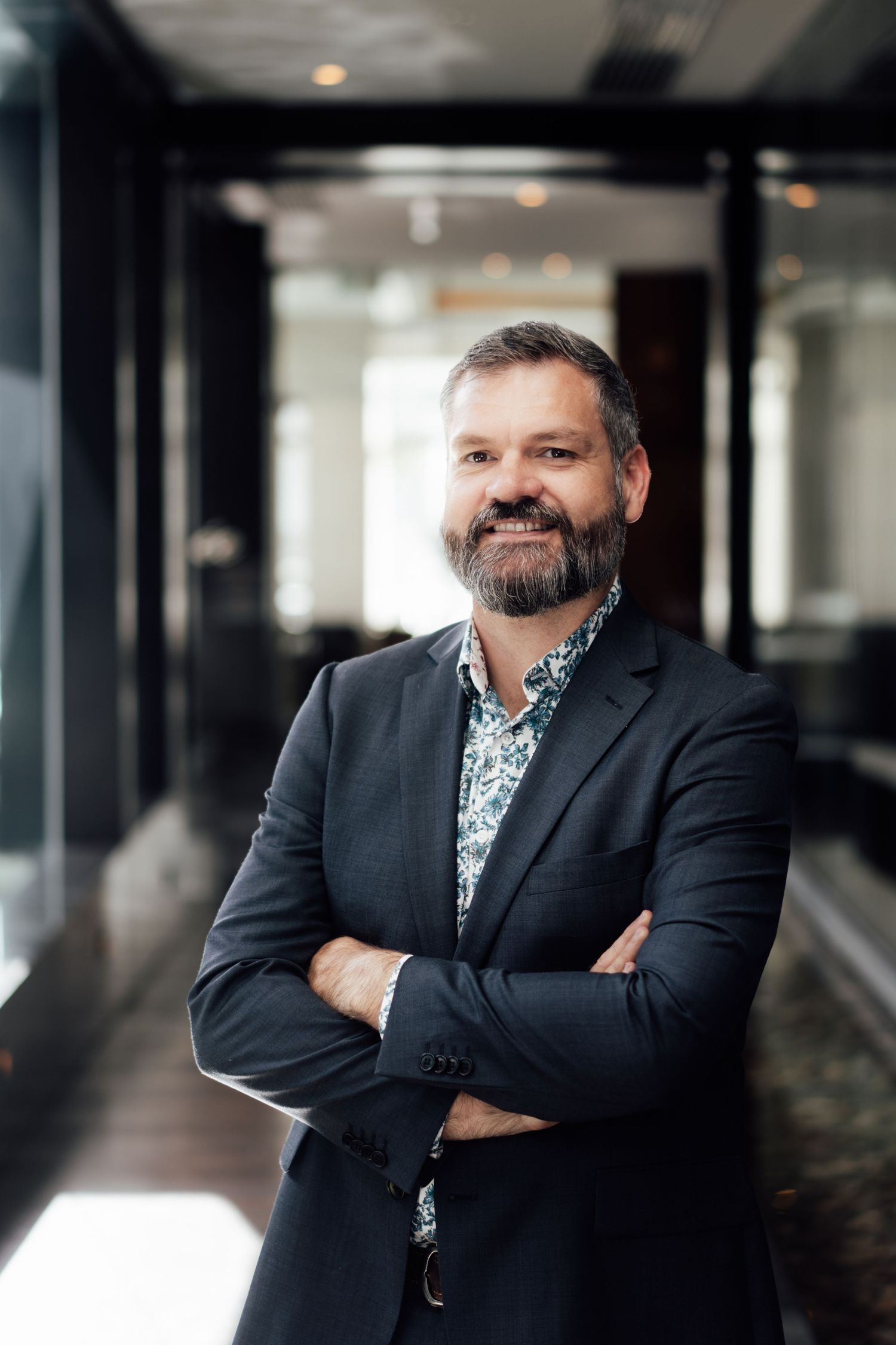 Smiling middle-aged bearded man in dark blazer and floral shirt standing with arms crossed indoors.