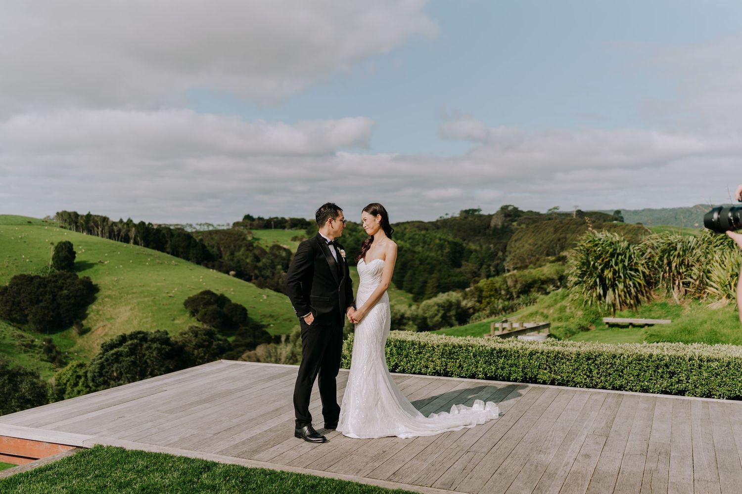 Newlyweds share intimate moment on wooden deck surrounded by lush green countryside.