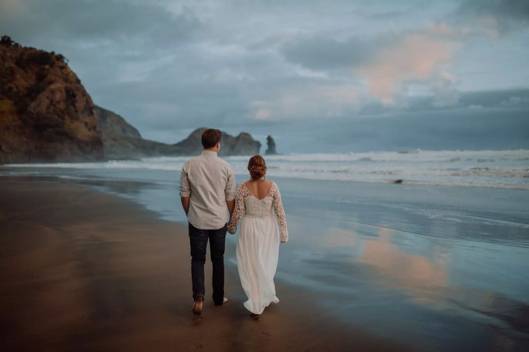 A couple walks along a misty beach at sunset with dramatic cliffs and moody skies in the background.