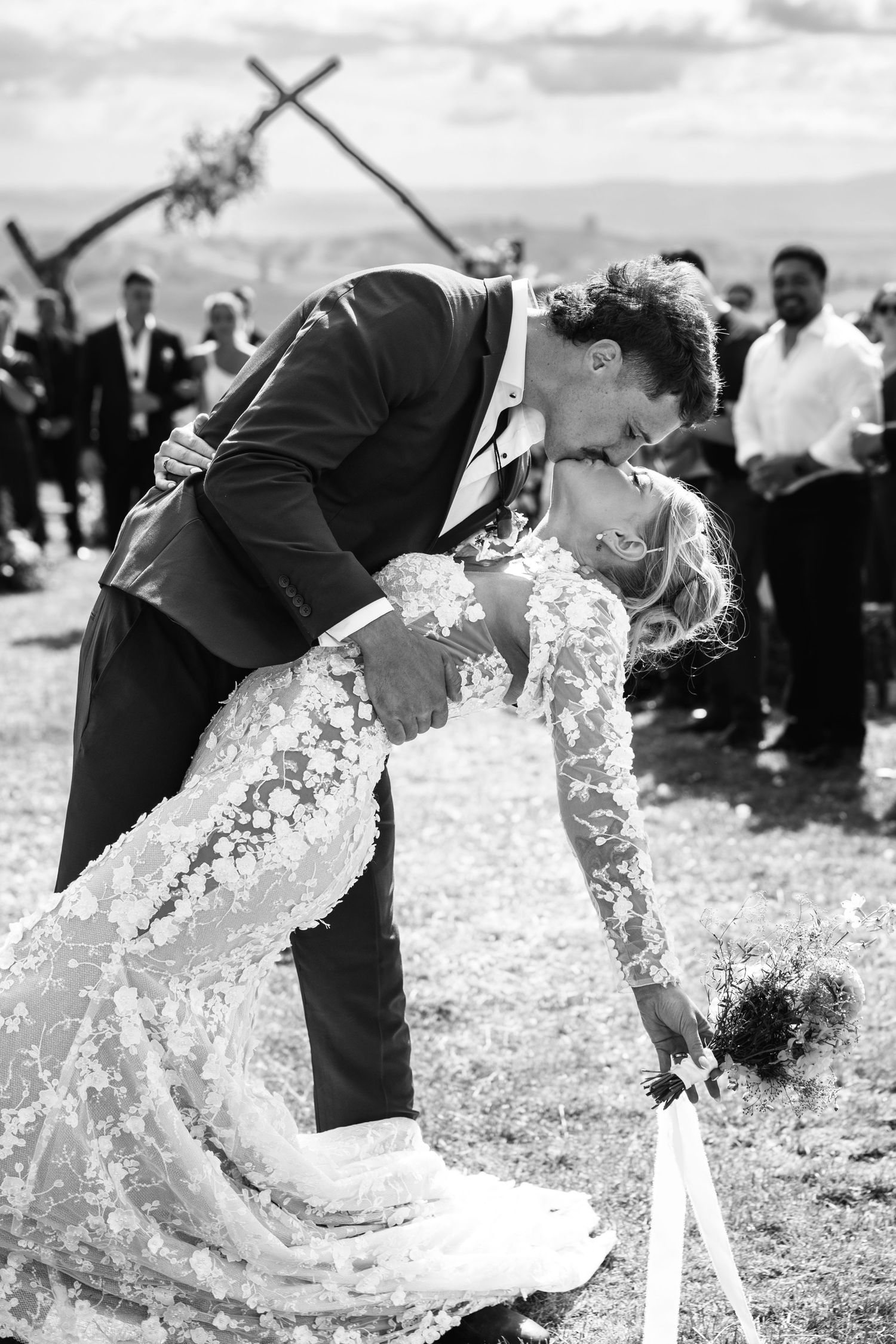Black and white photo of a groom dipping and kissing his bride in a lace wedding dress outdoors.