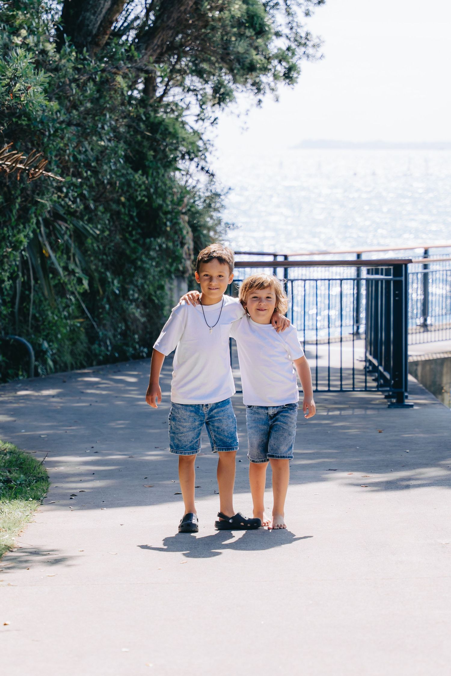 Two boys in white t-shirts and denim shorts stand together on a seaside path, smiling at the camera.