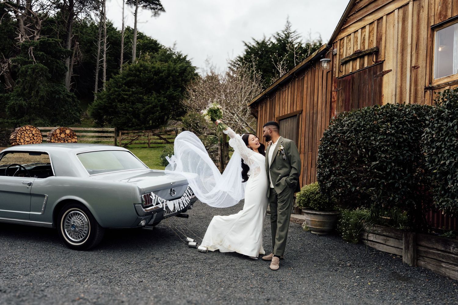 Bride and groom pose beside a classic mint Ford Mustang outside a rustic barn, veil flowing in the wind.