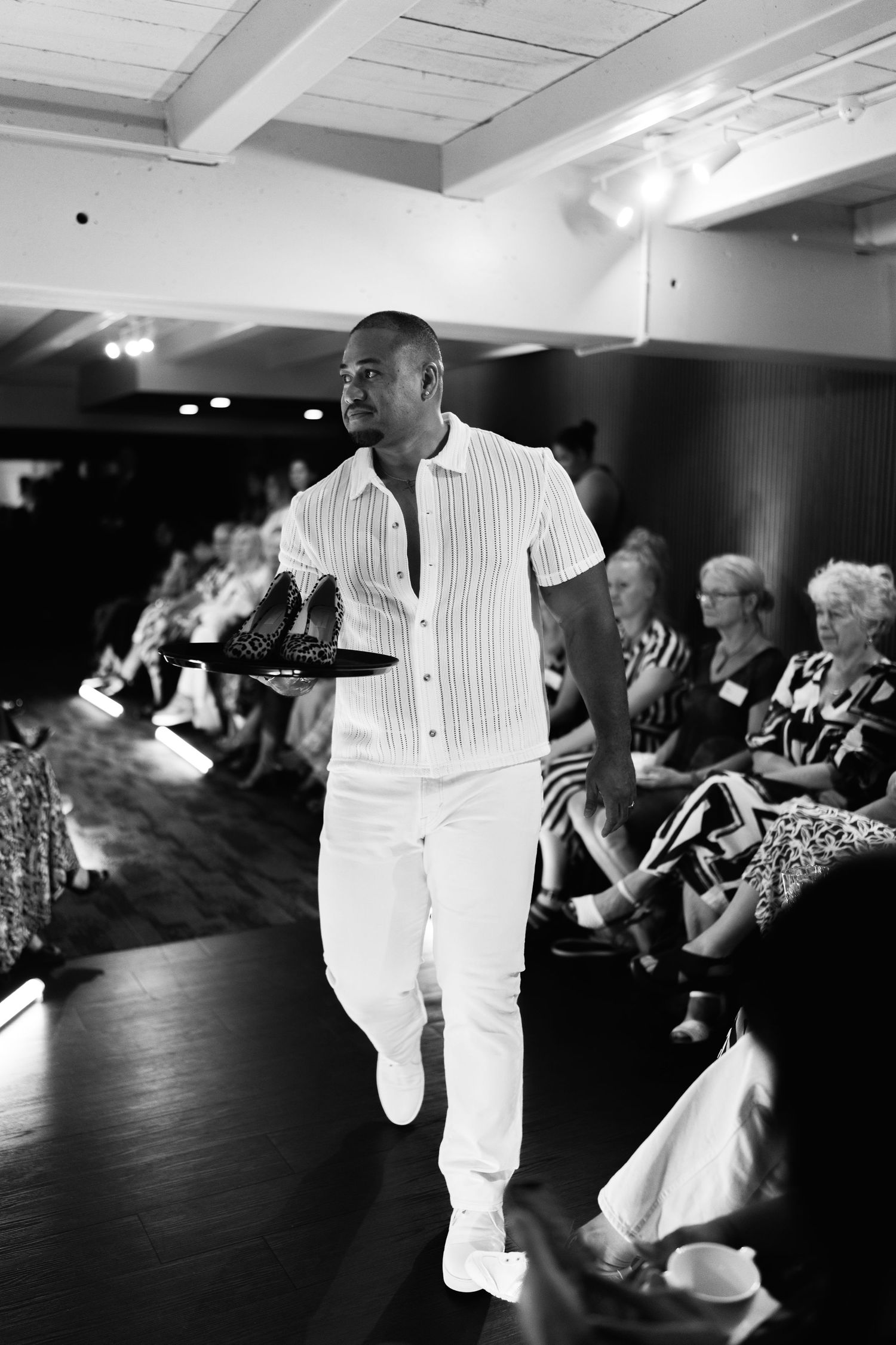 Black and white photo of a man in white outfit walking a fashion runway holding shoes, with seated audience watching.